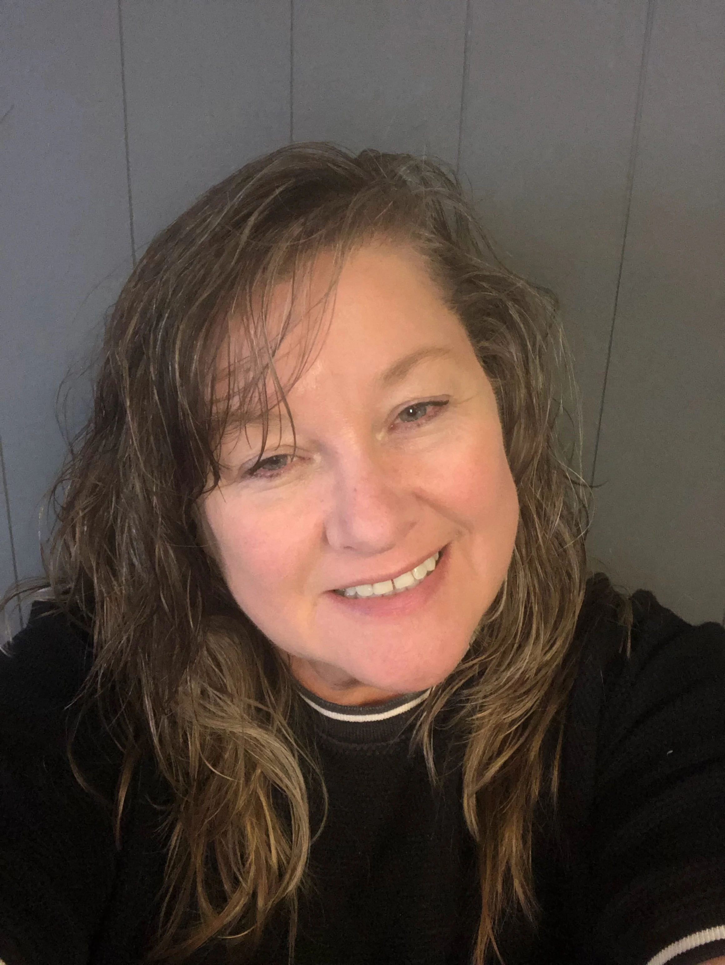 Close-up of a smiling woman with wavy hair, wearing a black top with white trim, against a gray wall background.