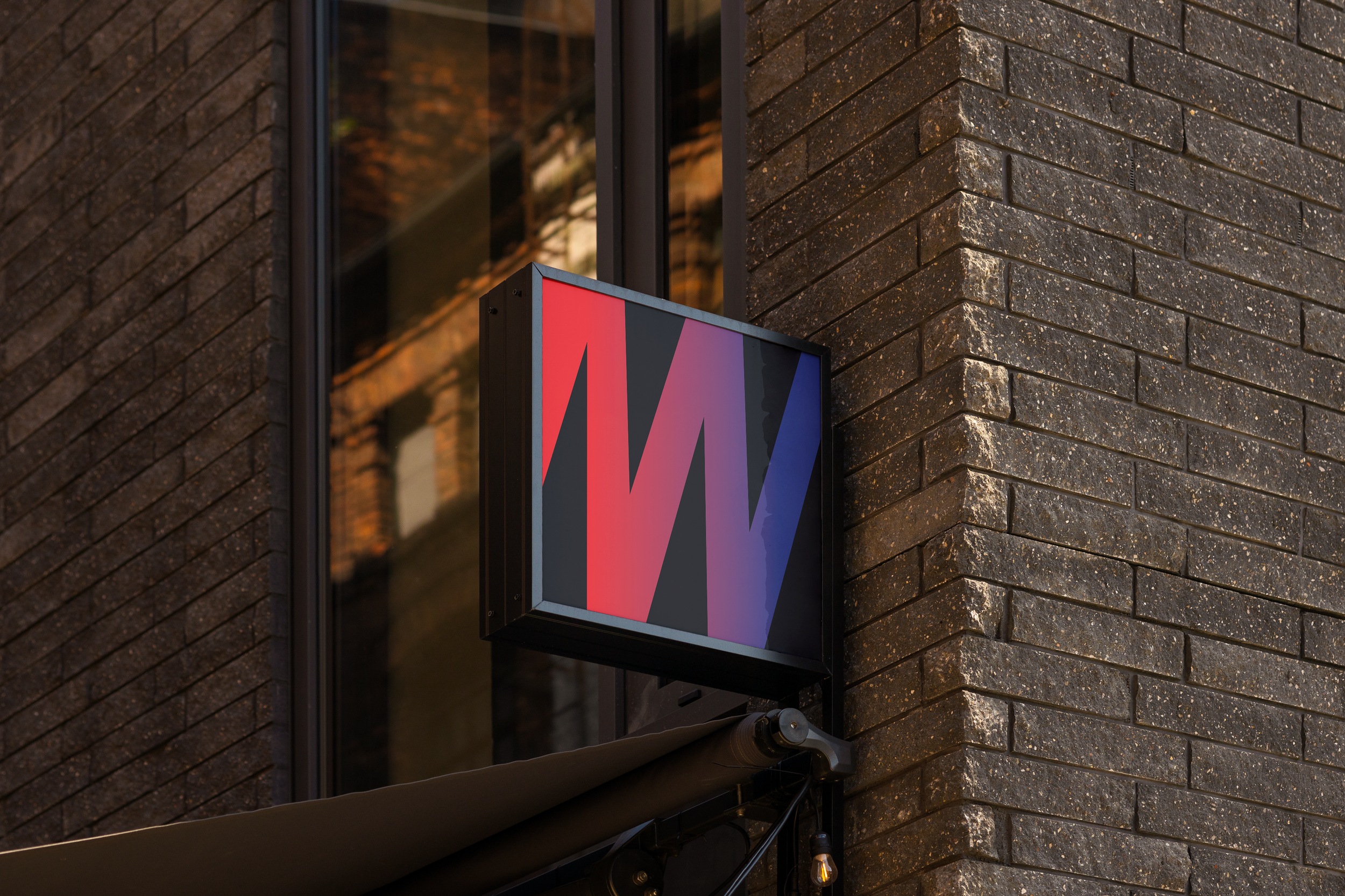 Close-up of a colorful rectangular logo sign with red, purple, and blue sections mounted on a dark brick building wall. The sign is illuminated and partially reflected in a window nearby.