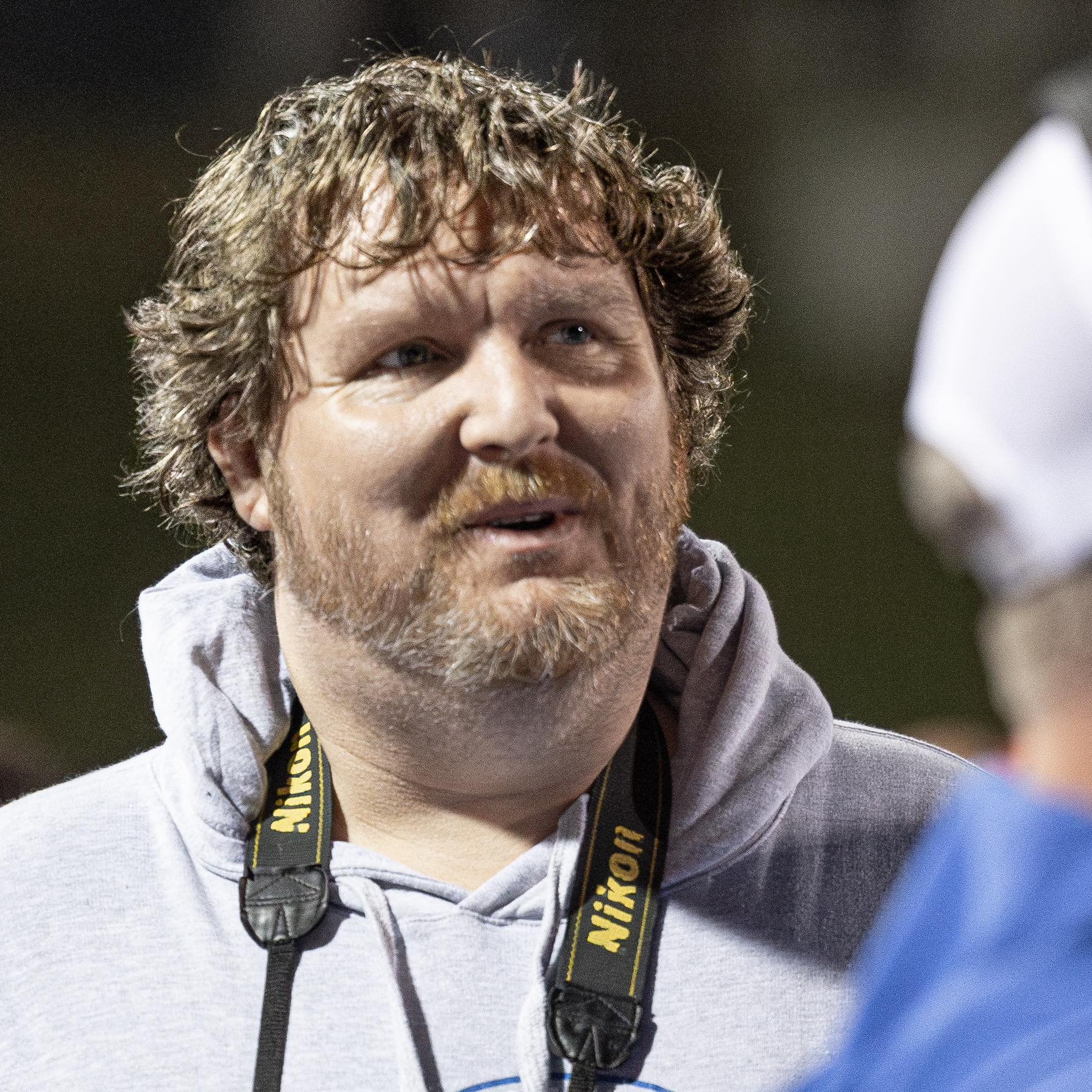 Close-up of a man with curly hair and a beard, wearing a hoodie and a camera strap, speaking or smiling, with blurred background.