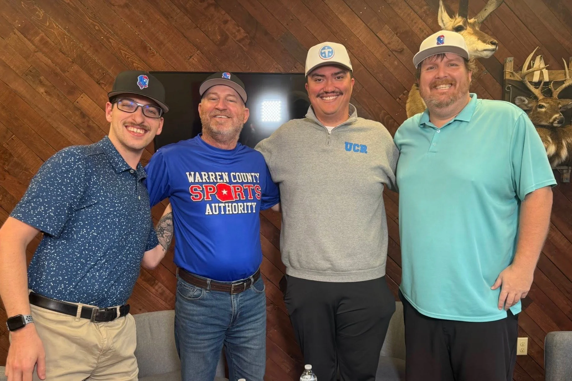 Four men standing together indoors with wood-paneled walls and deer heads mounted on the wall behind them. They are smiling and wearing casual clothing, including baseball caps.