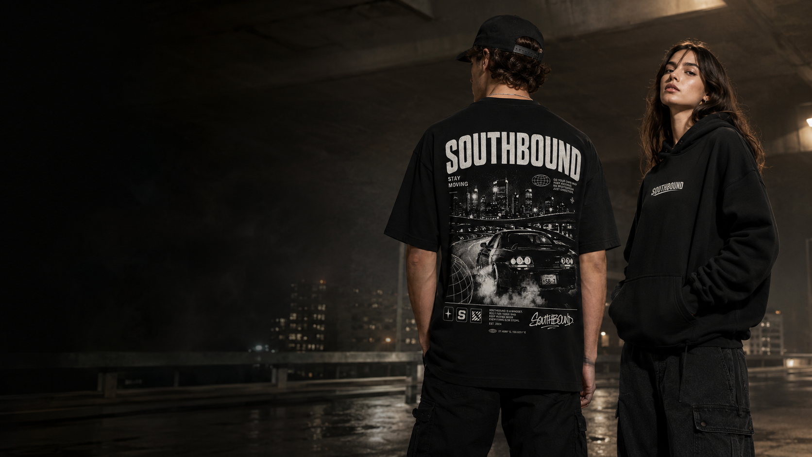 Two young people, one male and one female, standing under an urban overpass at night. The male is wearing a black cap and a large black t-shirt with graphics and the word 'SOUTHBOND'. The female is wearing a black hoodie with the word 'SOUTHBOND' on it. The background shows city lights and tall buildings.