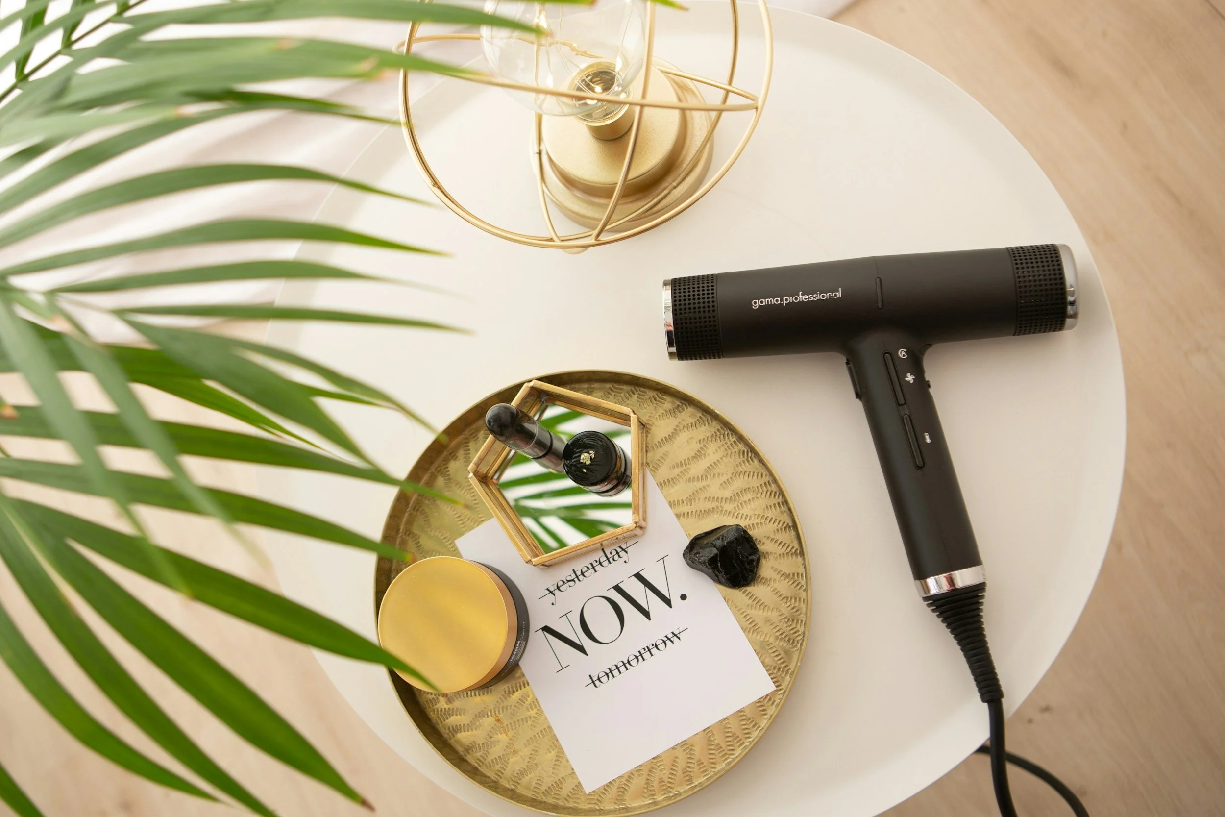 On a white table, a black handheld hair dryer, a glass lamp with metal frame, a small vase with black stones, a yellow container, a gold tray, and a magazine are arranged. Green leaves partially cover the view.