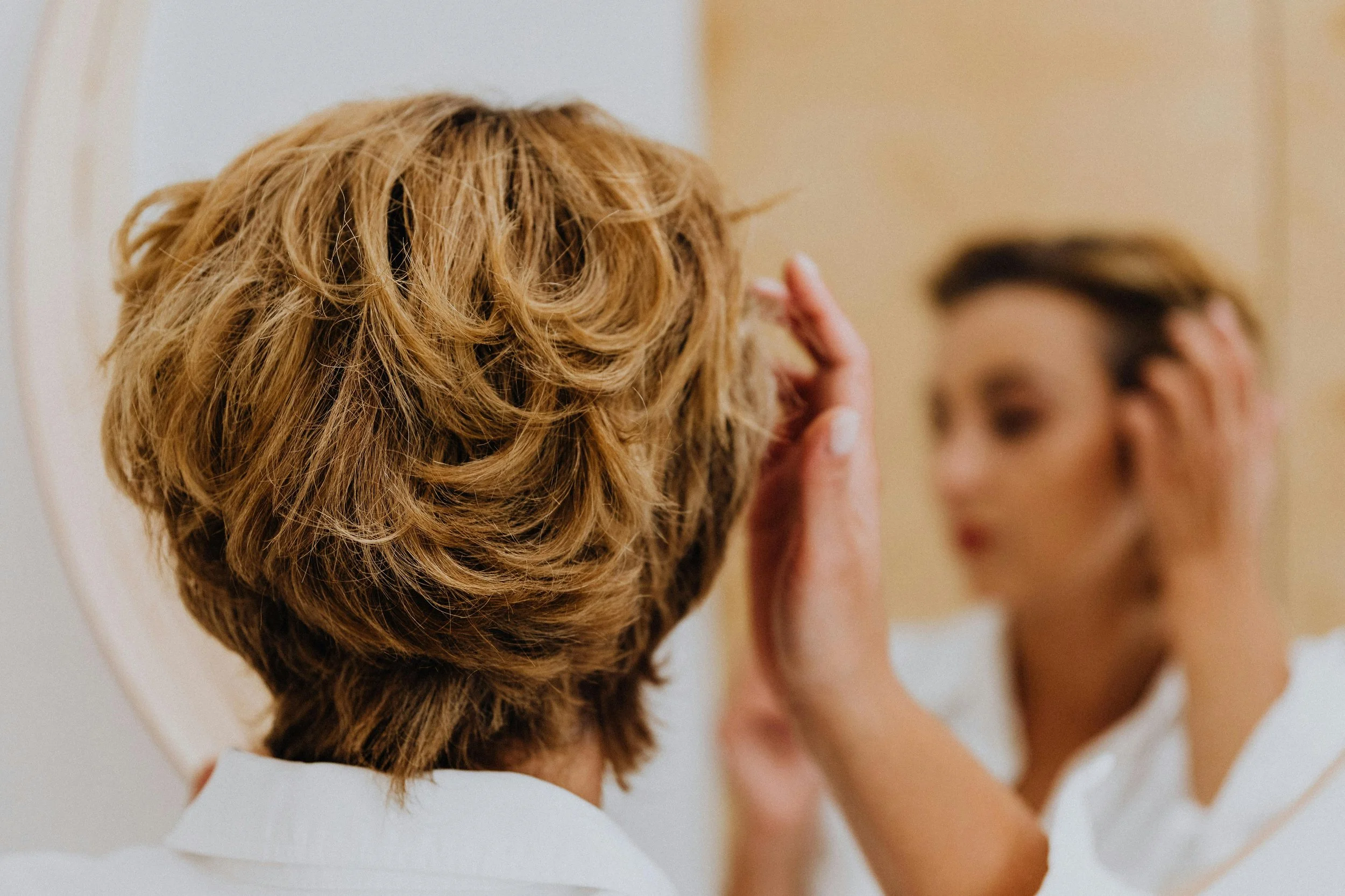 A person with short, slightly curly, blonde hair looking into a mirror. The person is touching their face with their right hand, and their facial features are blurred in the reflection. The background is neutral and soft in color.