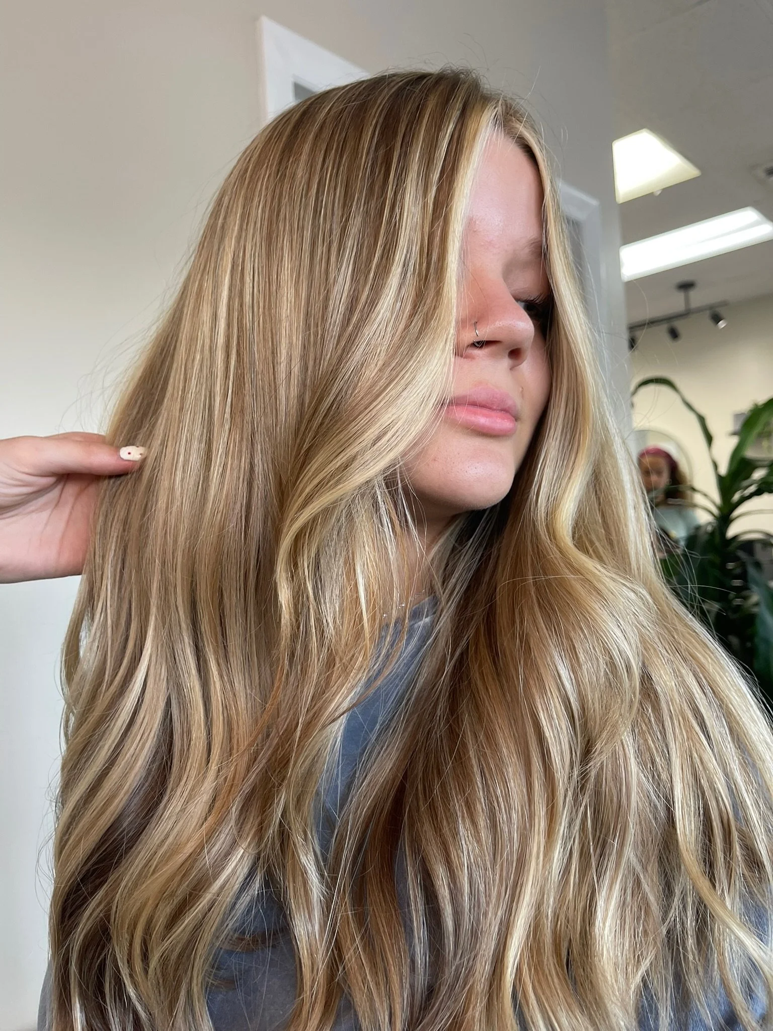 A woman with long, wavy blond hair, wearing a gray top, is seen in profile in a salon with bright lights and plants in the background.