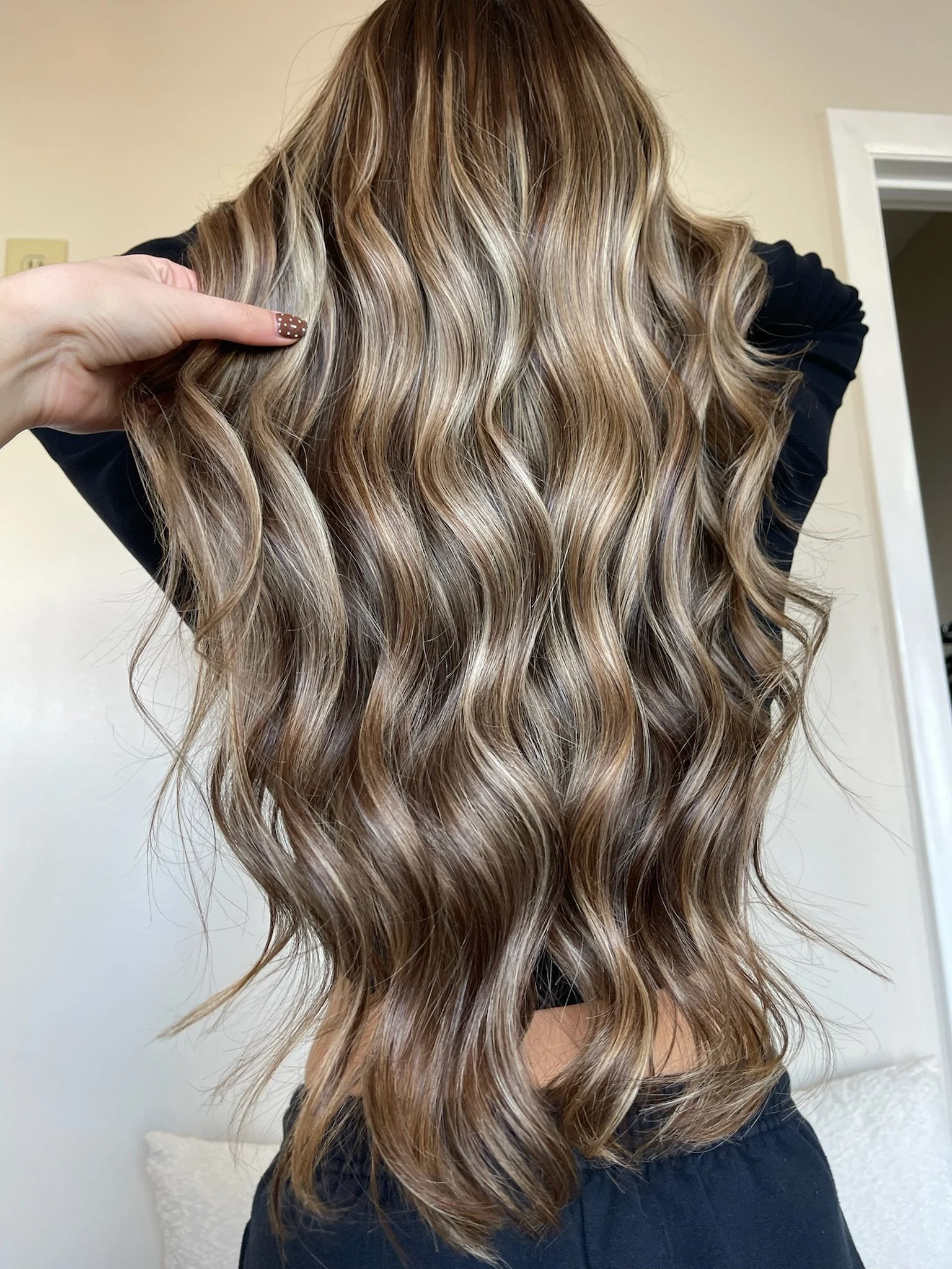 Person with long, wavy, blonde hair with darker roots, holding a section of hair. The hair is styled in loose waves with a shiny, healthy appearance, in front of a neutral-colored room.
