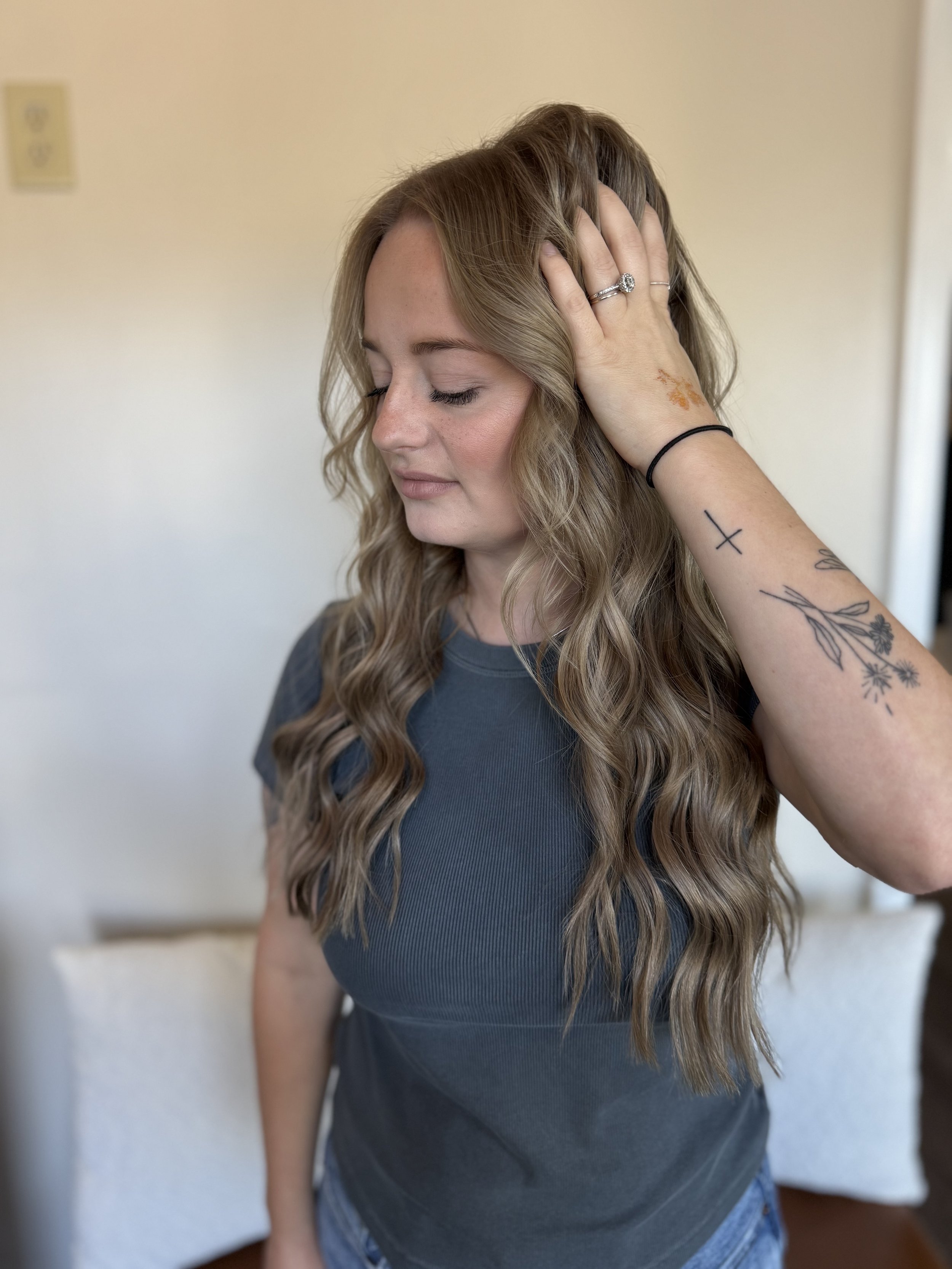 A woman with long, wavy blonde hair and freckles on her hand is holding her head. She has closed eyes and a calm expression. She is wearing rings, a black hair tie, and has tattoos on her forearm. The background is plain and light-colored.