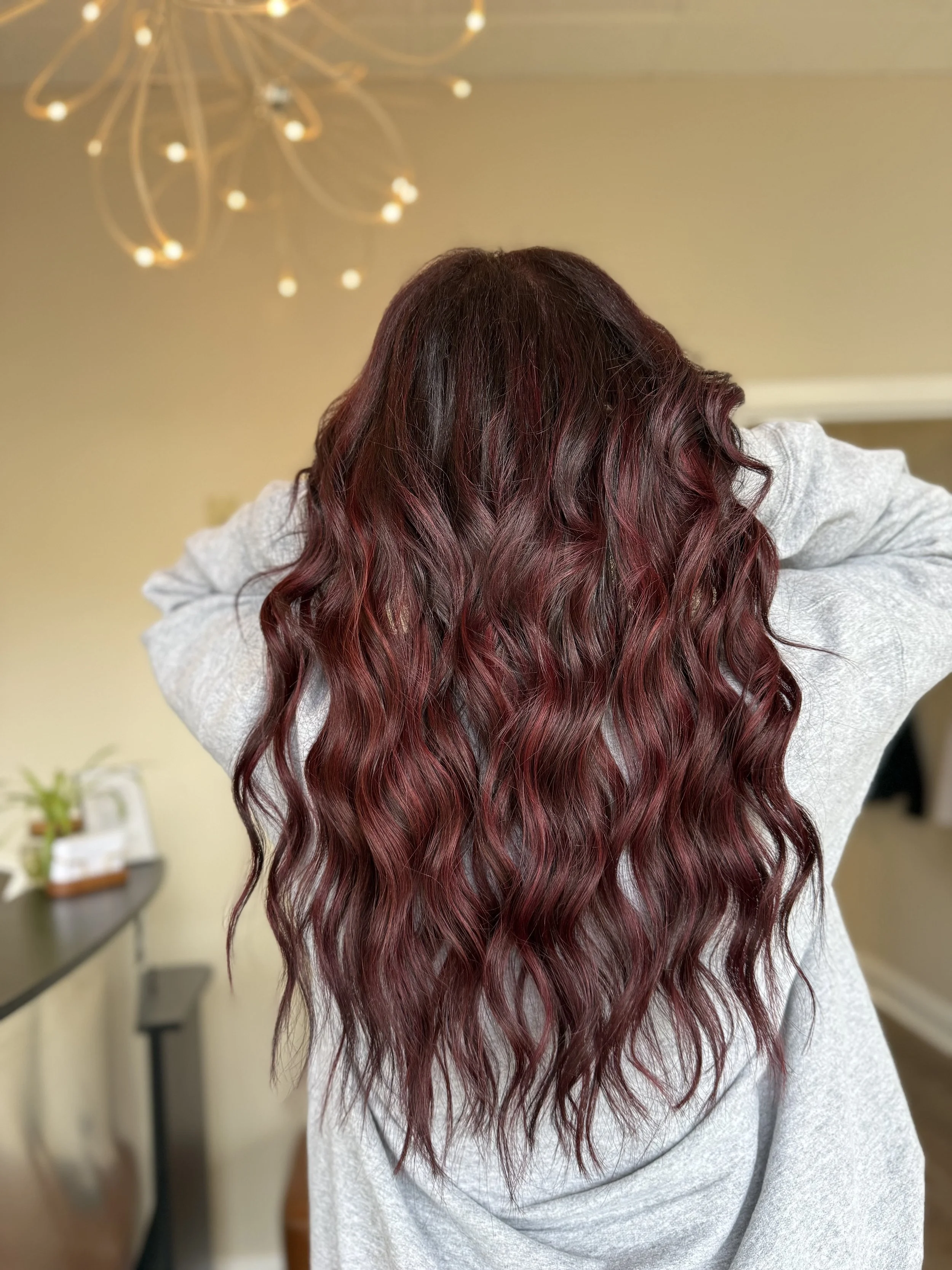 Back view of a person with long wavy hair dyed a deep red color, wearing a gray sweatshirt, with a blurred indoor background.