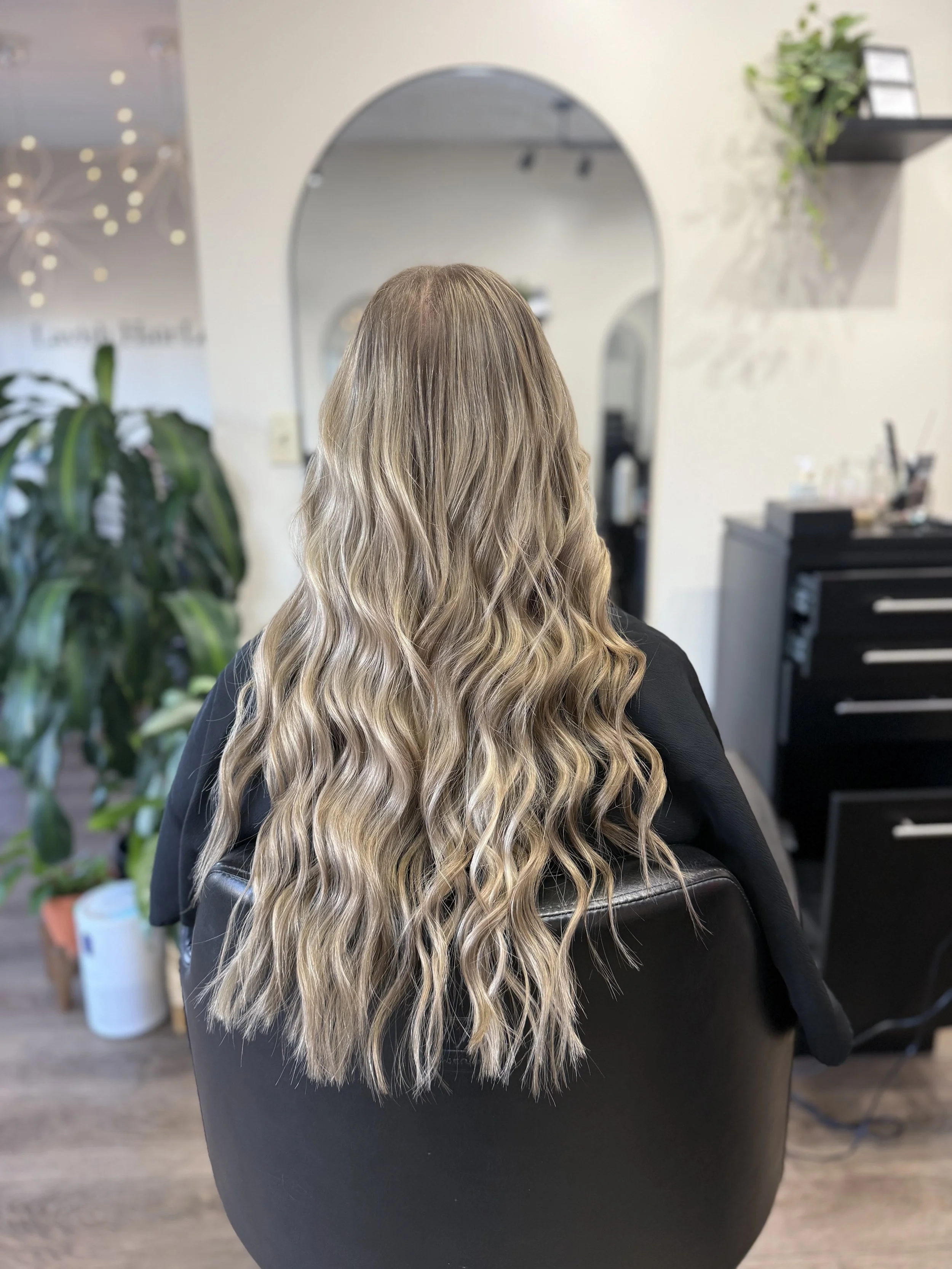 Hairdresser's chair with a woman sitting, showcasing long, wavy, blonde hair with highlights, in a salon setting with plants and shelves in the background.