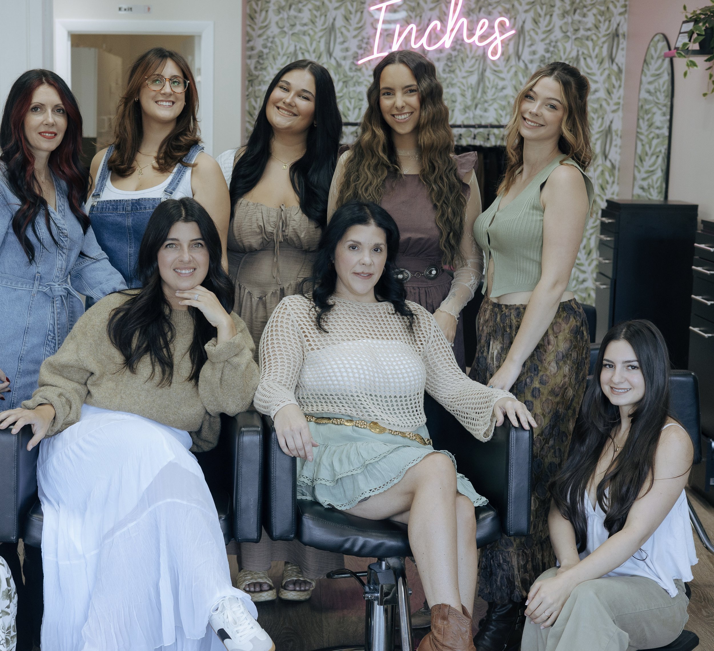 A group of women and young women smiling and posing for a photo inside a floral decorated room with neon pink sign that says "Inches."