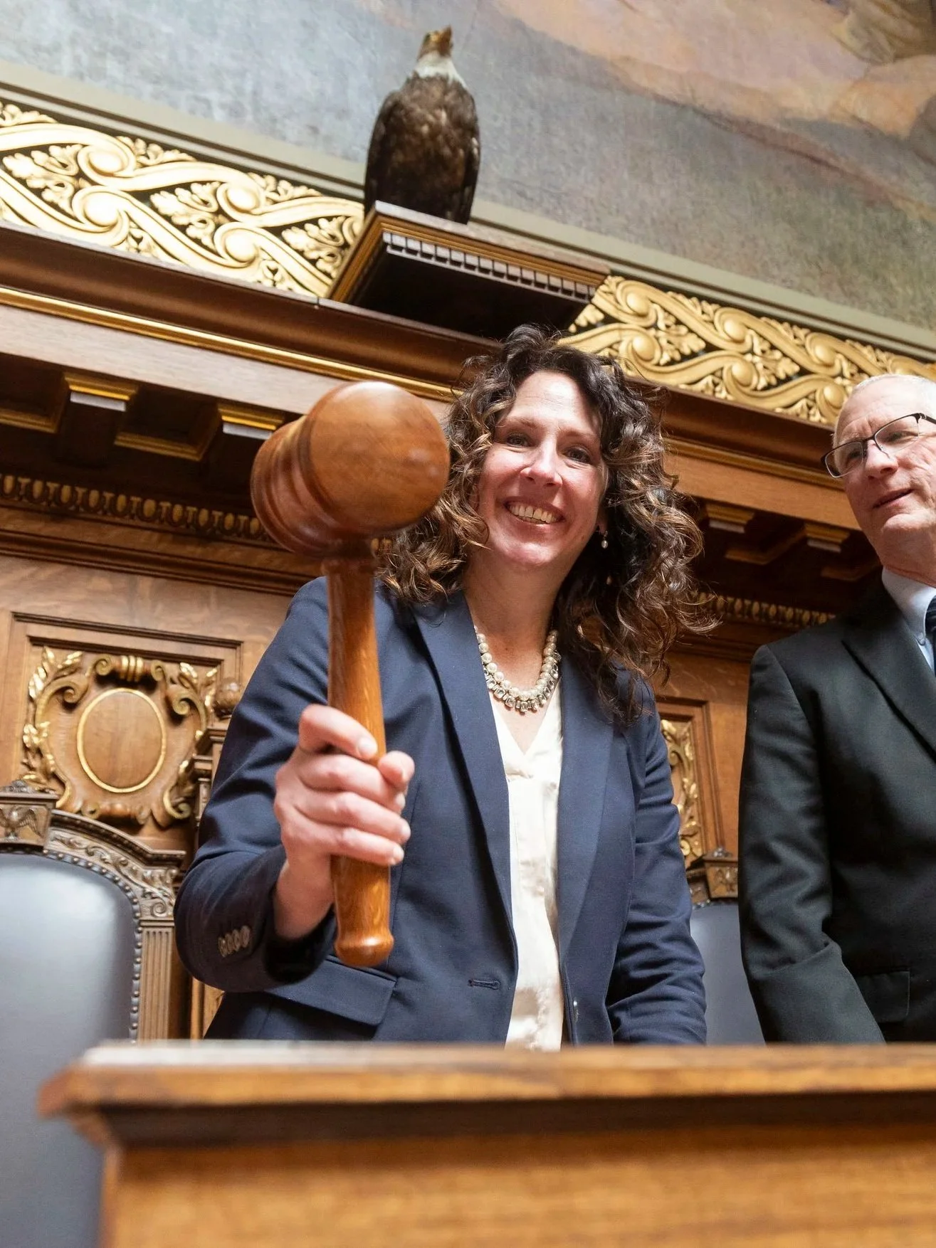 A woman in a blue blazer holding a wooden gavel in a courtroom or formal setting with ornate woodwork and decorative elements.