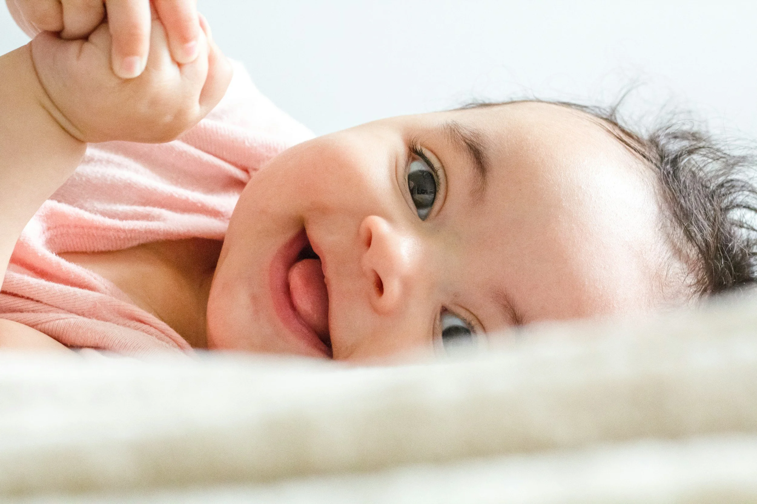 A close-up photo of a young girl lying down, with her head turned to the side. She has light skin, big blue eyes, and dark curly hair. She is wearing a pink and white striped shirt and slightly smiling.