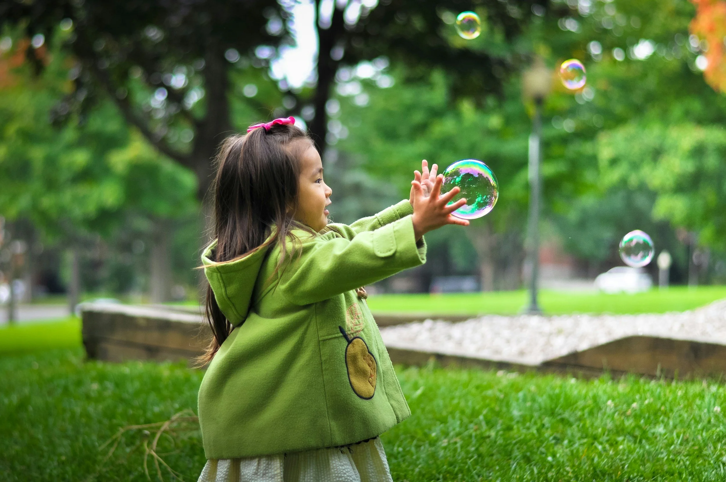 A young girl with long brown hair wearing a green hoodie with a teddy bear design reaching out to touch soap bubbles floating in the air in a park with green trees in the background.