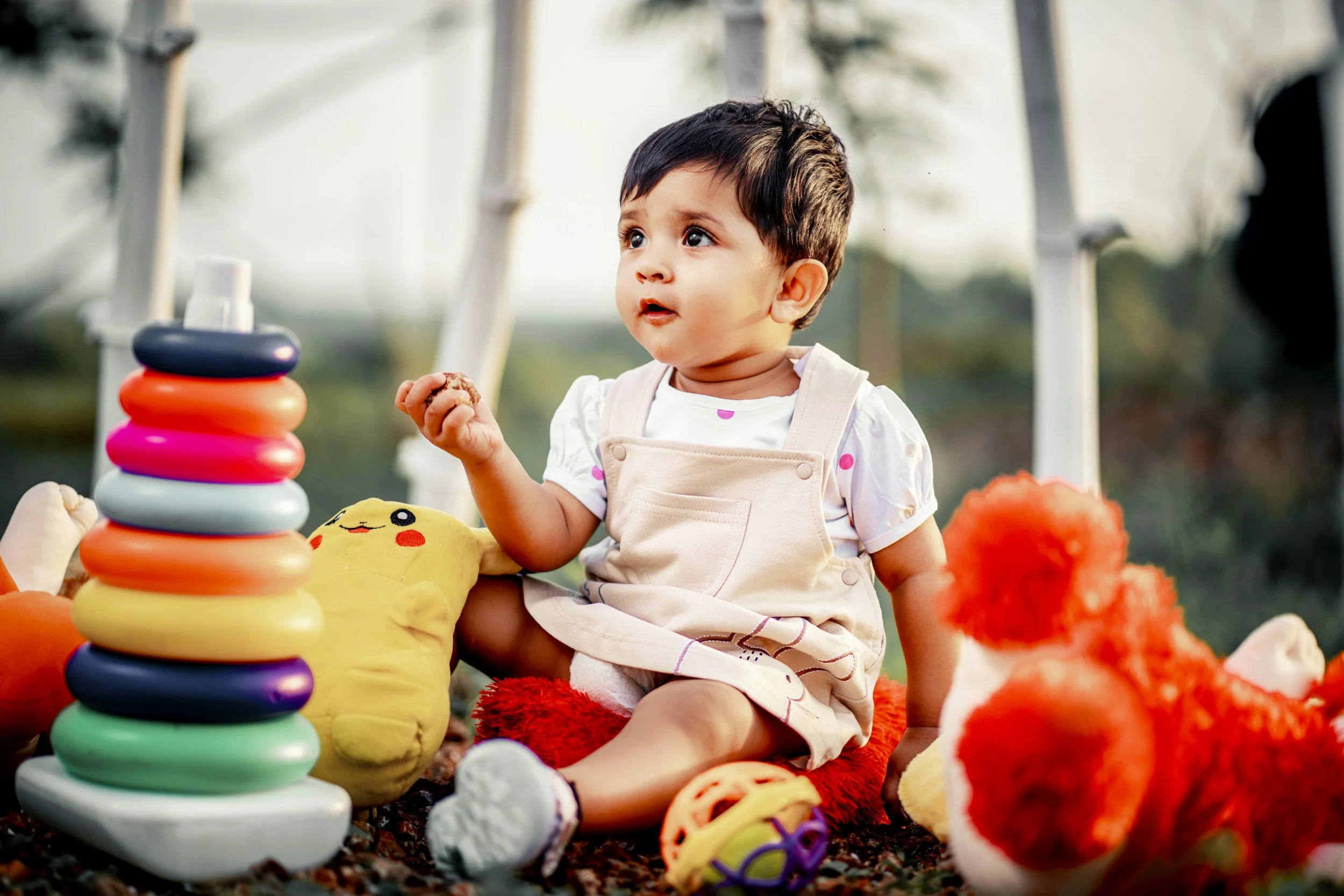 Young boy sitting outdoors surrounded by colorful plush toys and a stacking ring toy, looking attentive and curious.