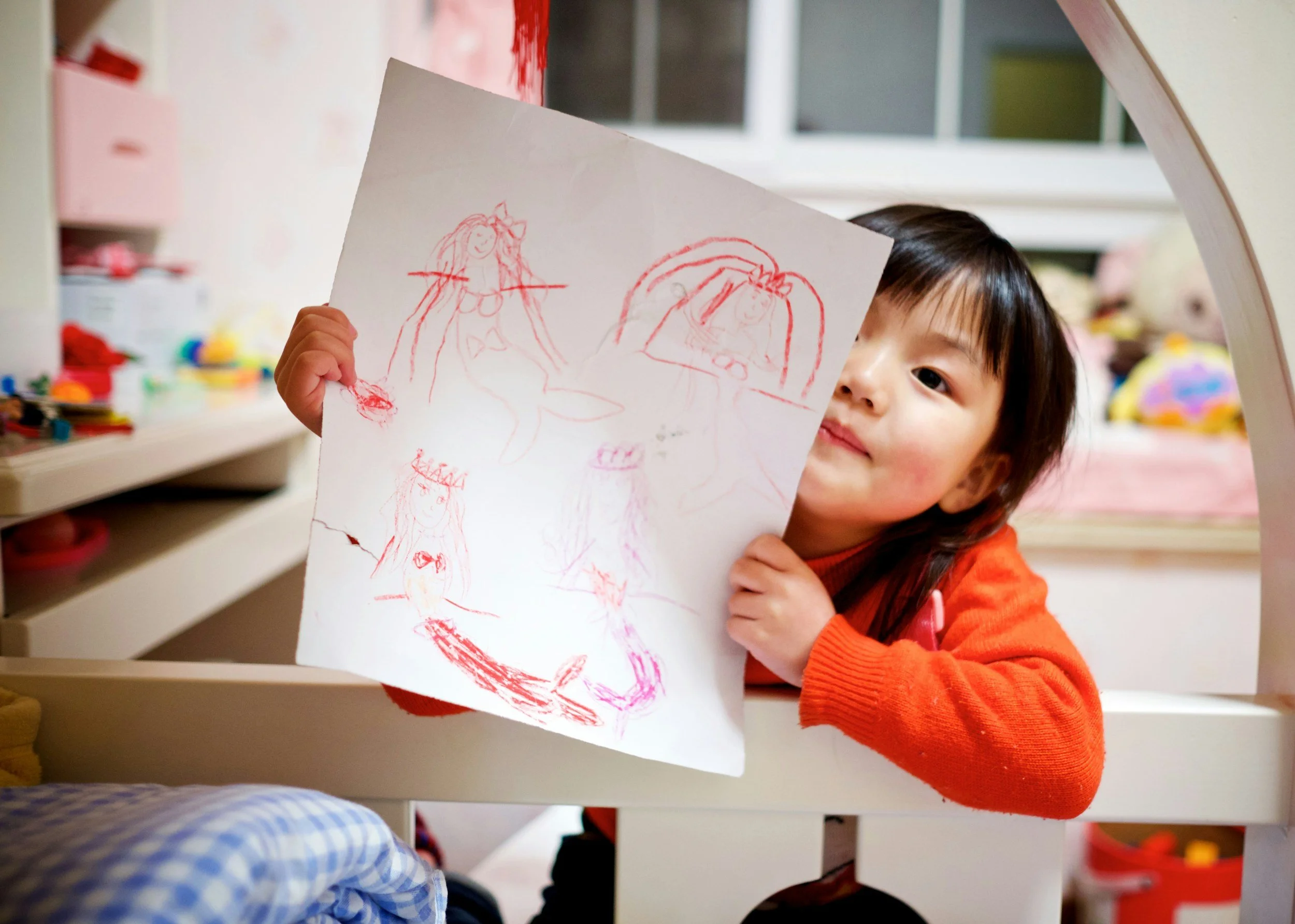 A young girl with dark hair and an orange sweater showing a drawing of a princess on a piece of paper.