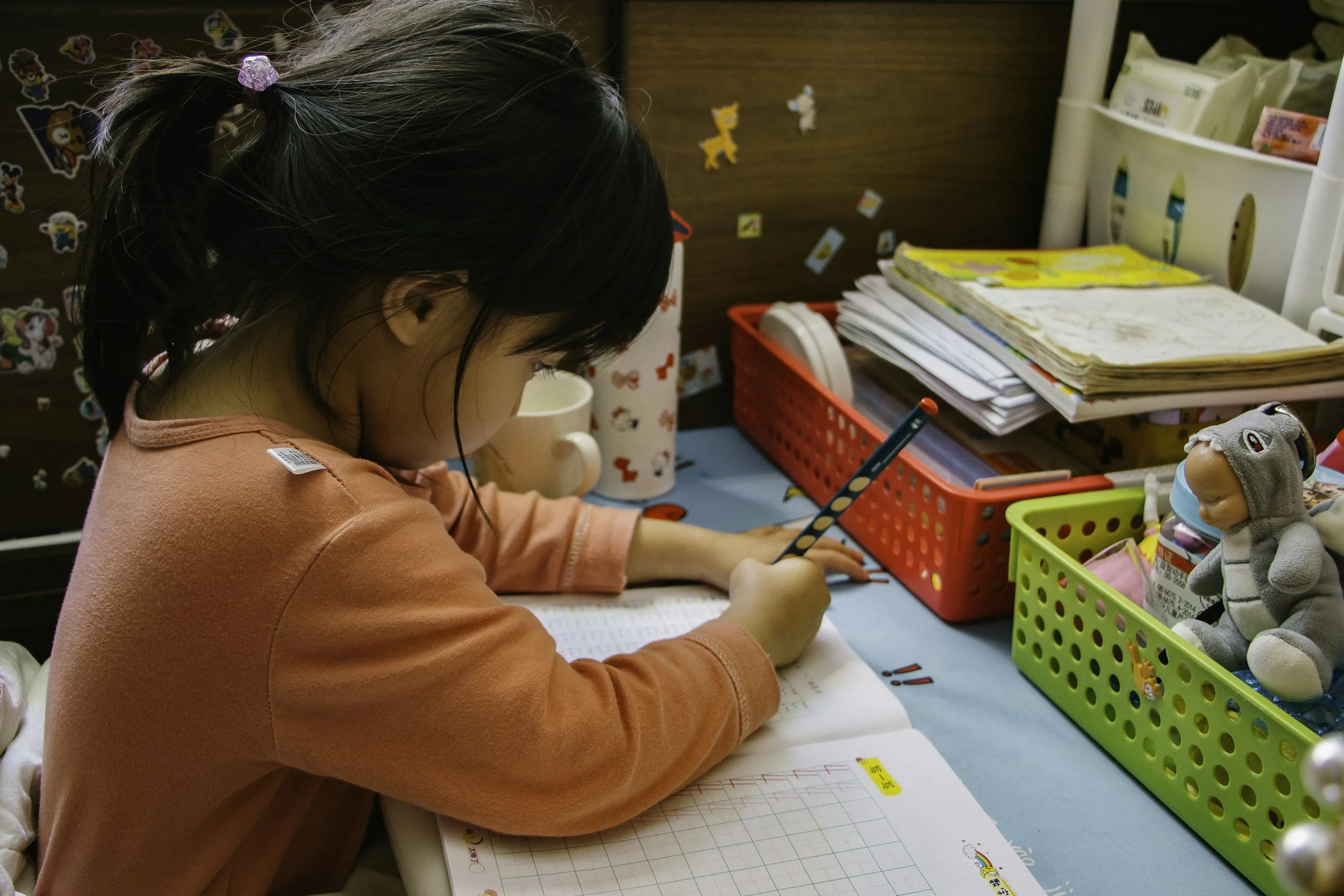A young girl with dark hair in a pink long-sleeve shirt, sitting at a desk, working on schoolwork or drawing with a pencil. There are various notebooks, papers, and colored markers organized in colorful baskets on the desk. The background has a wall decorated with stickers of cartoon characters and animals, and a shelf with more school supplies and a doll.