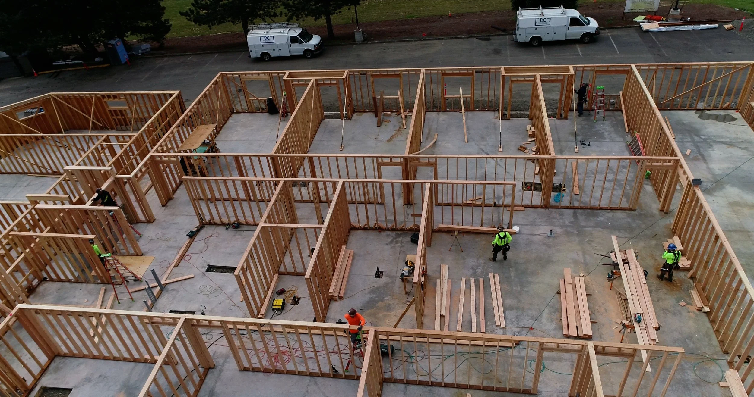 Construction site with wooden framing for building walls, workers wearing safety vests and helmets, and several utility vehicles parked nearby.