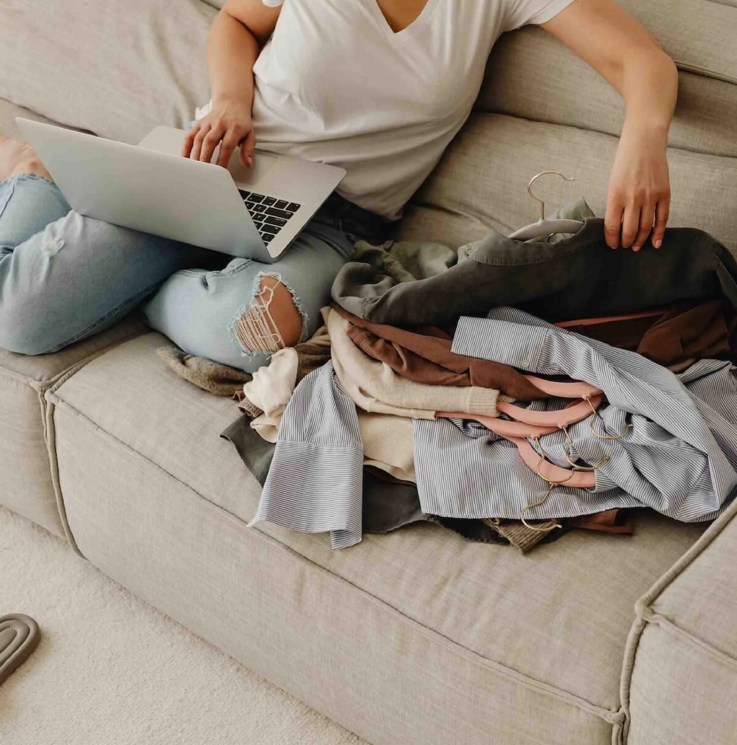 Person sorting through a pile of clothes on a sofa with a laptop open