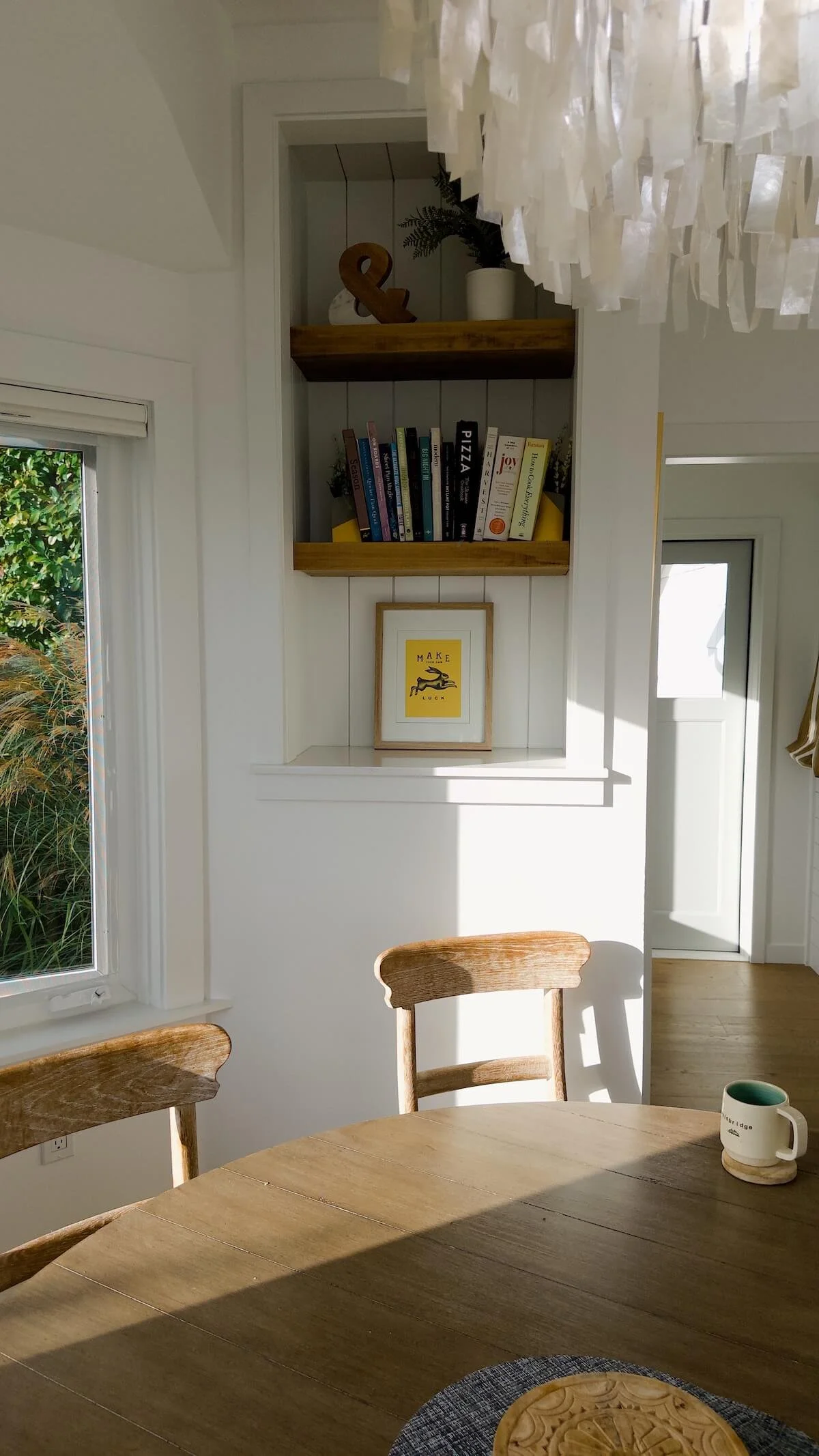 Sunlit, uncluttered kitchen nook with cookbooks on timber shelves and a round wooden table, from a post on how to organise a pantry