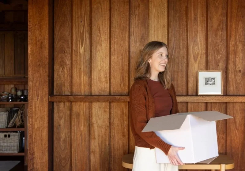 Smiling woman in a rust cardigan carrying an open white donation box past wooden walls, part of learning how to organize a pantry