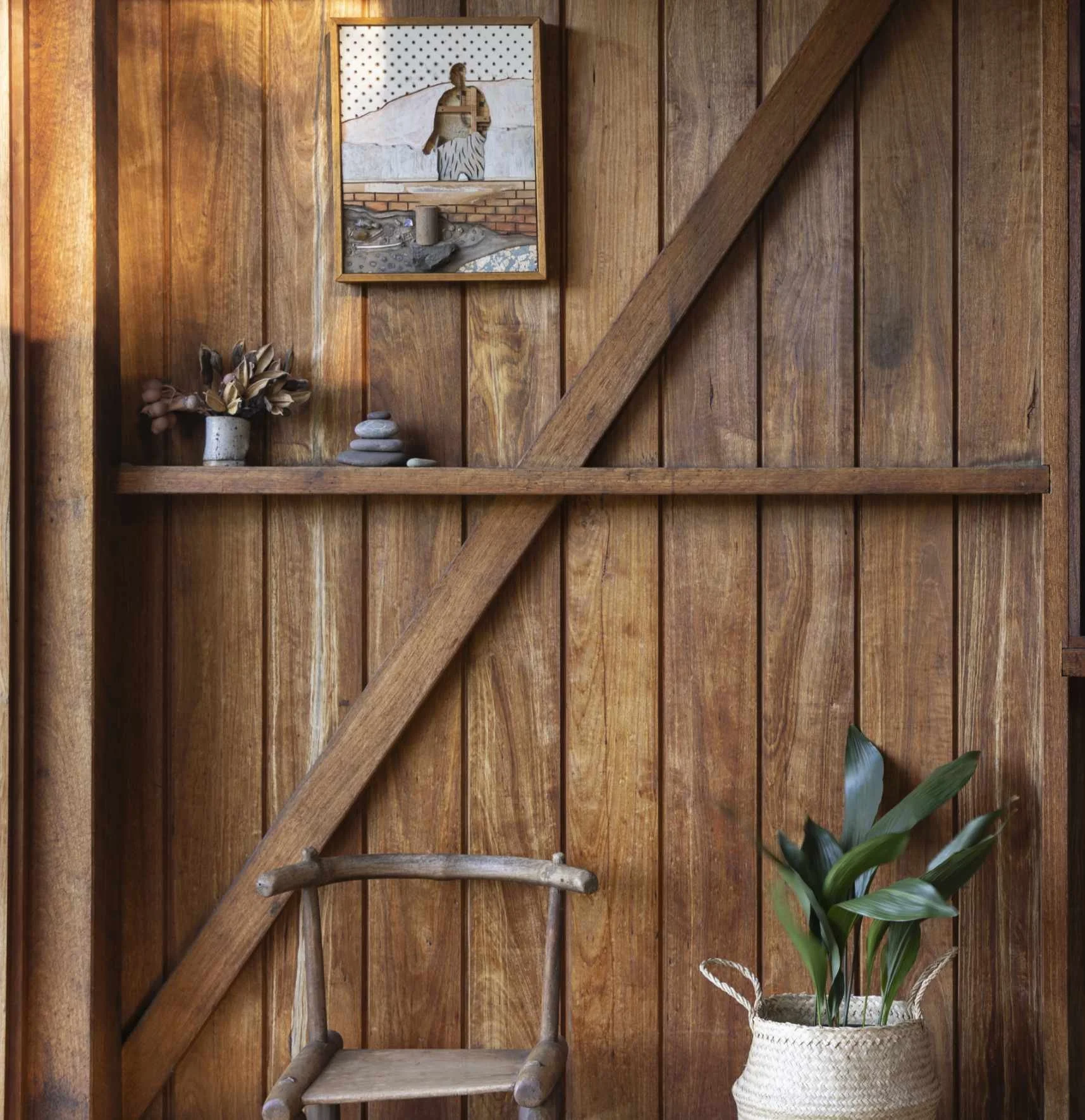 Rustic timber interior, diagonal wooden beams, a shelf with stones and a small artwork, a wooden chair, and a large leafy plant in a woven basket.