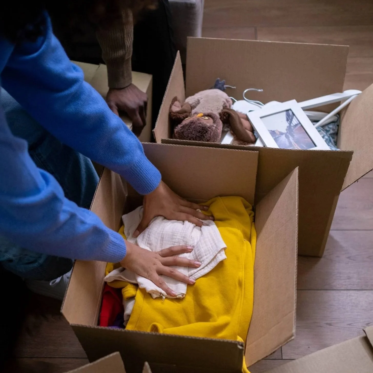 Two people with brown skin place unwanted household items into boxes for donations, for Hello Space Online Decluttering Coach. We can only see their hands and arms.
