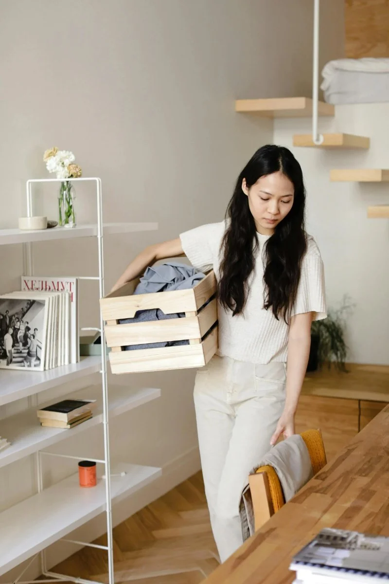 Woman carrying a wooden crate of clothes past white shelves, learning how to declutter your home