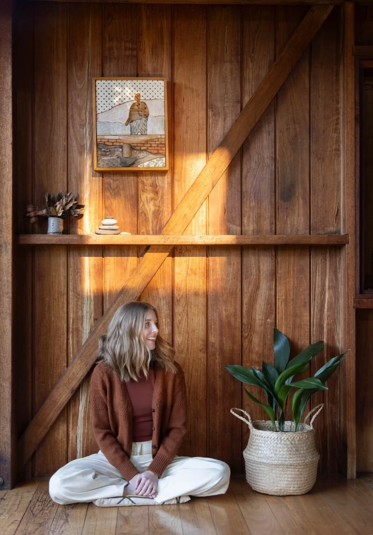 Dr Bea Bobbin, Canberra home organiser and decluttering coach, sitting cross-legged in a warm timber space surrounded by plants and handmade objects