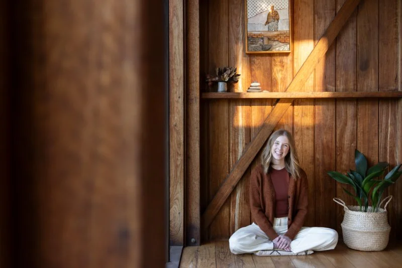 Dr Bea Bobbin sitting cross-legged and smiling in a warm, sunlit wooden room, part of the Hello Space guide to what is decluttering.
