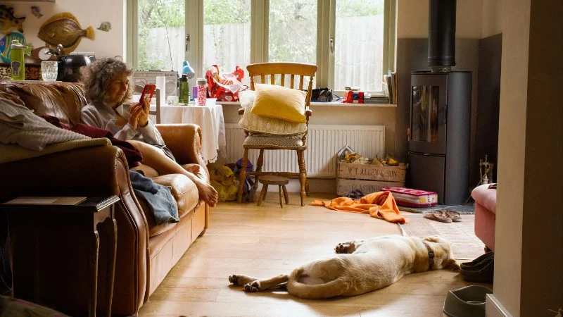 A woman relaxes with her dog in a lived-in living room full of everyday clutter, toys, boxes, and clothes, the kind of scene that makes people ask what is decluttering and where to start.