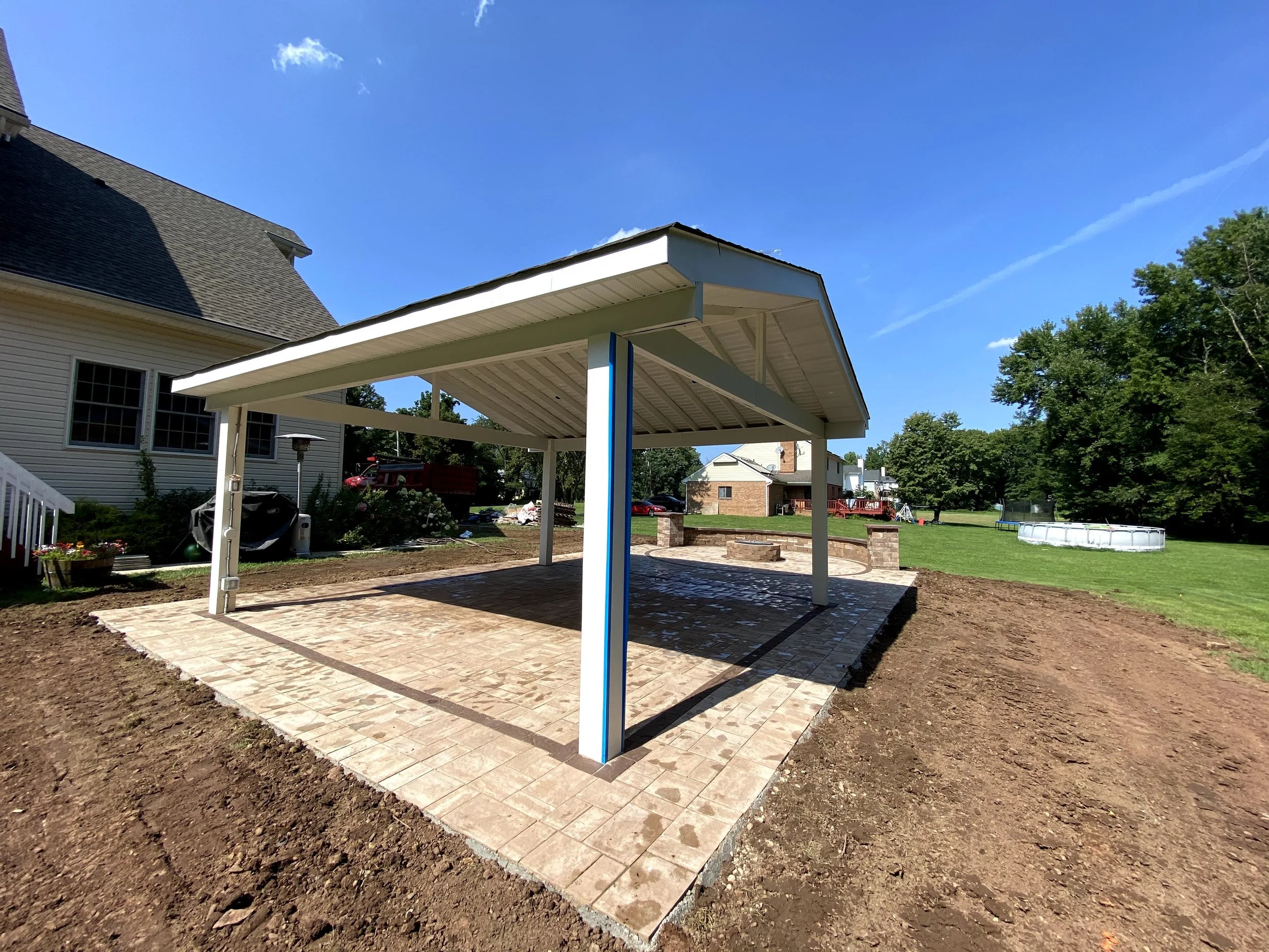Backyard with newly laid brick patio under a white metal pergola, surrounded by soil and a white house on the left with windows, green trees, and a blue sky with clouds.