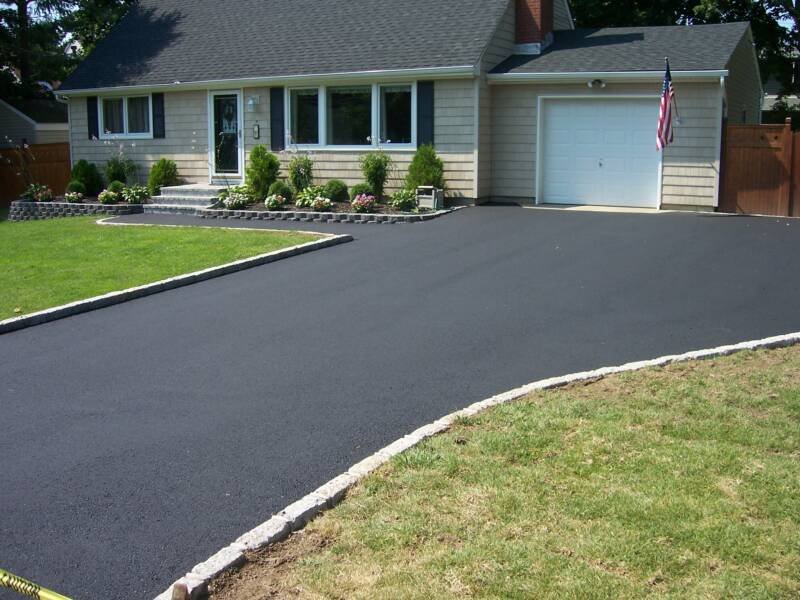 Newly paved black asphalt driveway leading to a suburban house with a garage and American flag, with a well-maintained lawn and garden beds.