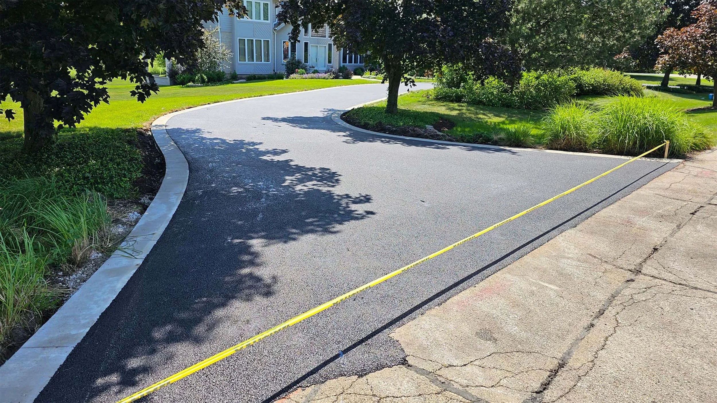 Newly paved asphalt driveway with a yellow caution tape outlining the edge, surrounded by green grass and trees in a residential neighborhood.