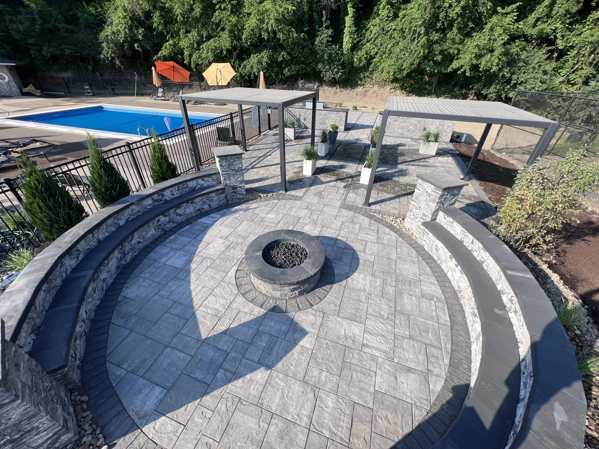 An outdoor pool area with a blue rectangular swimming pool, surrounded by a patio with gray stone pavers. There are two shade structures with modern metal frames and covered with fabric. Several potted plants and chairs with umbrellas are around the pool area. A hillside with dense green trees borders the scene.