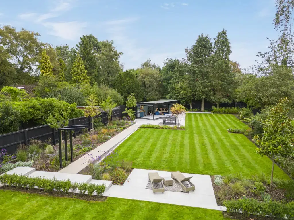 A spacious backyard with well-maintained green lawn, concrete pathways, a sitting area with outdoor furniture, and a garden with various plants and trees enclosed by a black fence.