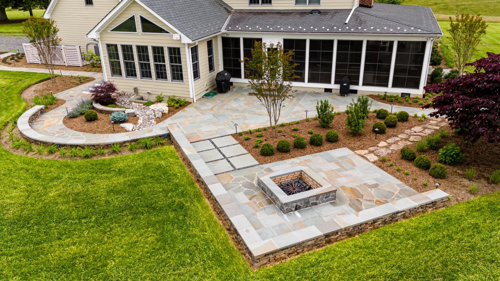 A spacious backyard patio with stone tiles, landscaped garden beds, small trees, shrubs, and a built-in fire pit surrounded by a low stone wall near a house with screened porch.