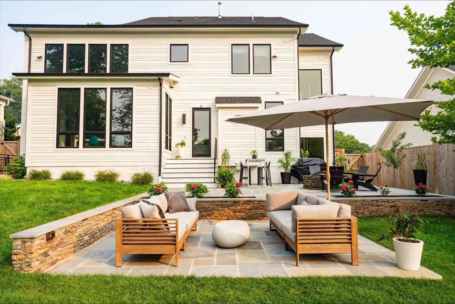 Backyard with a patio, outdoor seating, umbrella, grill, and a two-story white house with large windows.