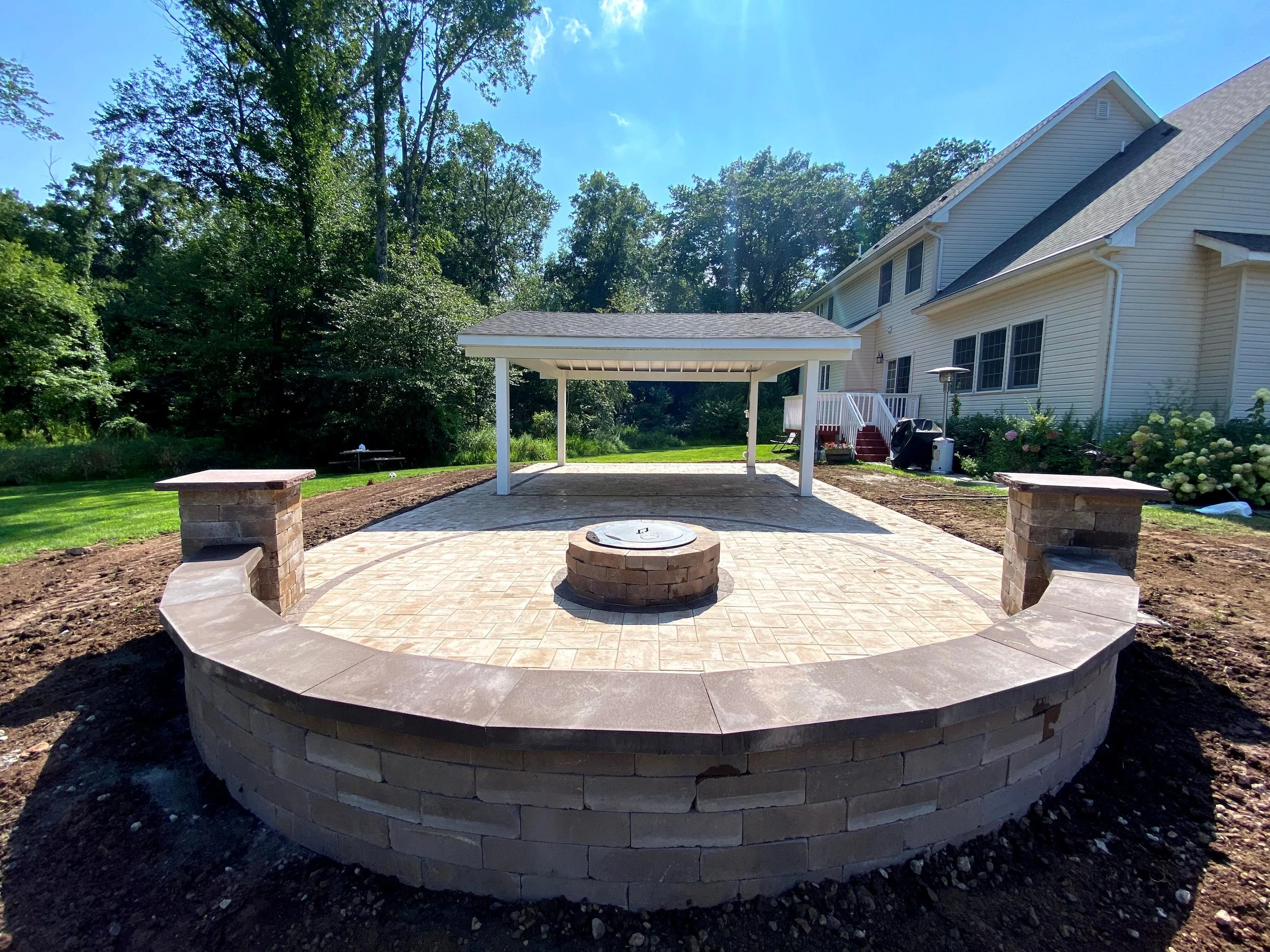 A backyard with a raised circular stone fire pit and a patio area with a pavilion, surrounded by green grass and trees, with a two-story house with beige siding in the background.