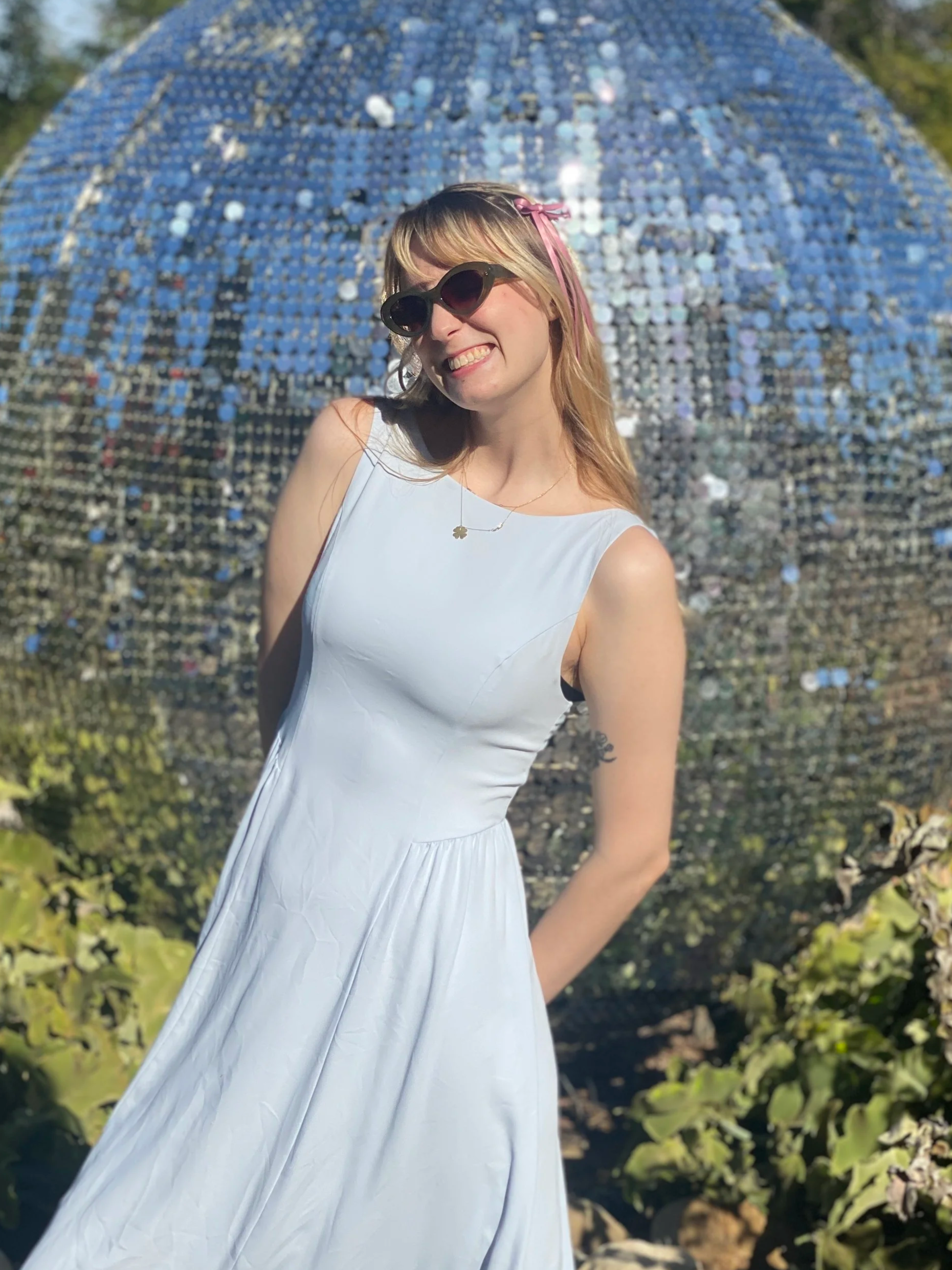 A young woman with blonde hair, wearing sunglasses, a light-colored sleeveless dress, a necklace, and a pink ribbon in her hair, standing outdoors in front of a large reflective spherical sculpture made of small mirrored tiles.