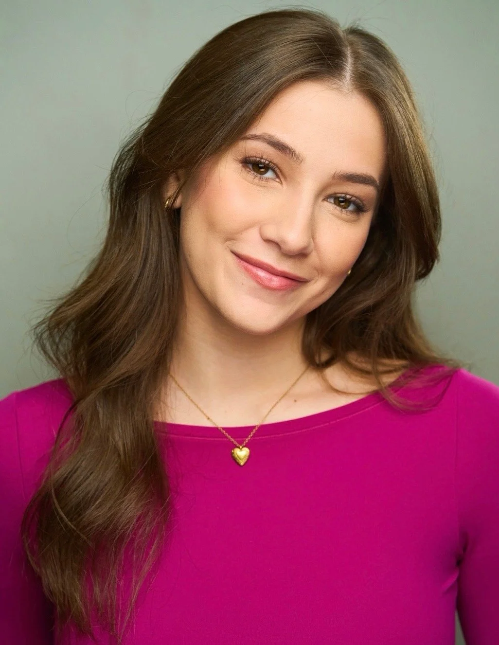 Portrait of a smiling young woman with long brown hair, wearing a pink top and a gold heart necklace, against a plain background.
