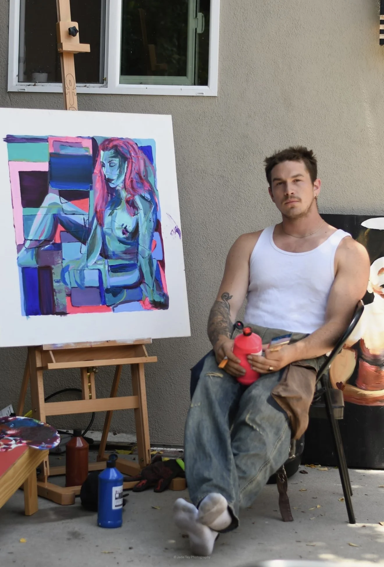 A young man with tattoos wearing a white tank top and distressed jeans sits outside on a black chair next to a colorful abstract painting of a woman with pink hair on an easel. Around him are art supplies, bottles of paint, and a red water bottle. The scene appears to be a backyard or outdoor art space with a beige wall and a window in the background.