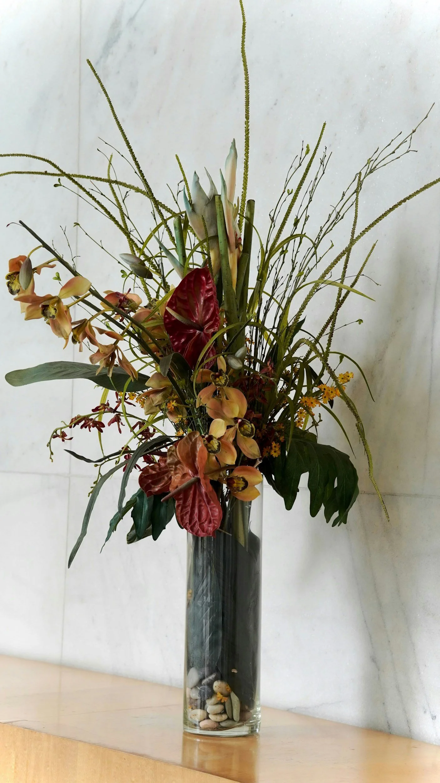 A tall glass vase with a floral arrangement of various flowers and plants, including orchids, large green leaves, and long, thin stems with small green buds and seed pods, set on a wooden surface against a plain white wall.