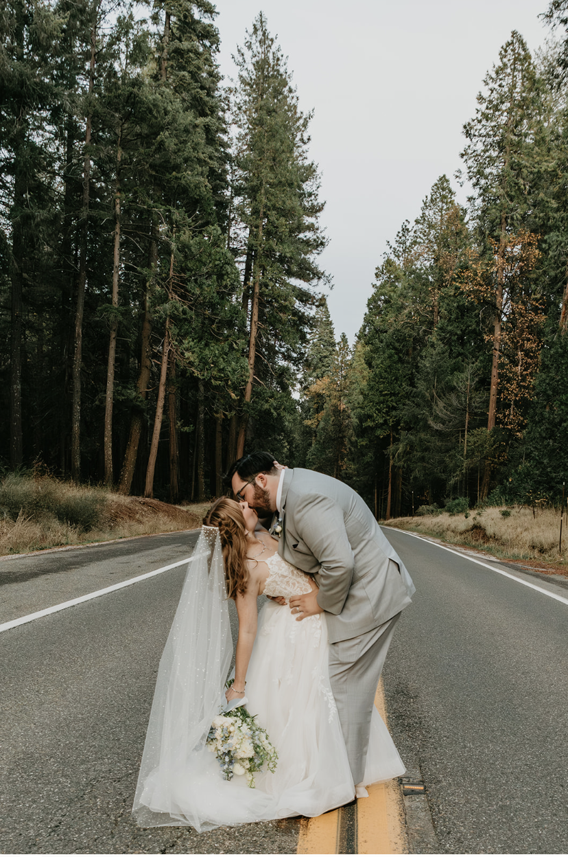 A bride and groom share a kiss in the middle of a two-lane road surrounded by tall trees on a cloudy day.