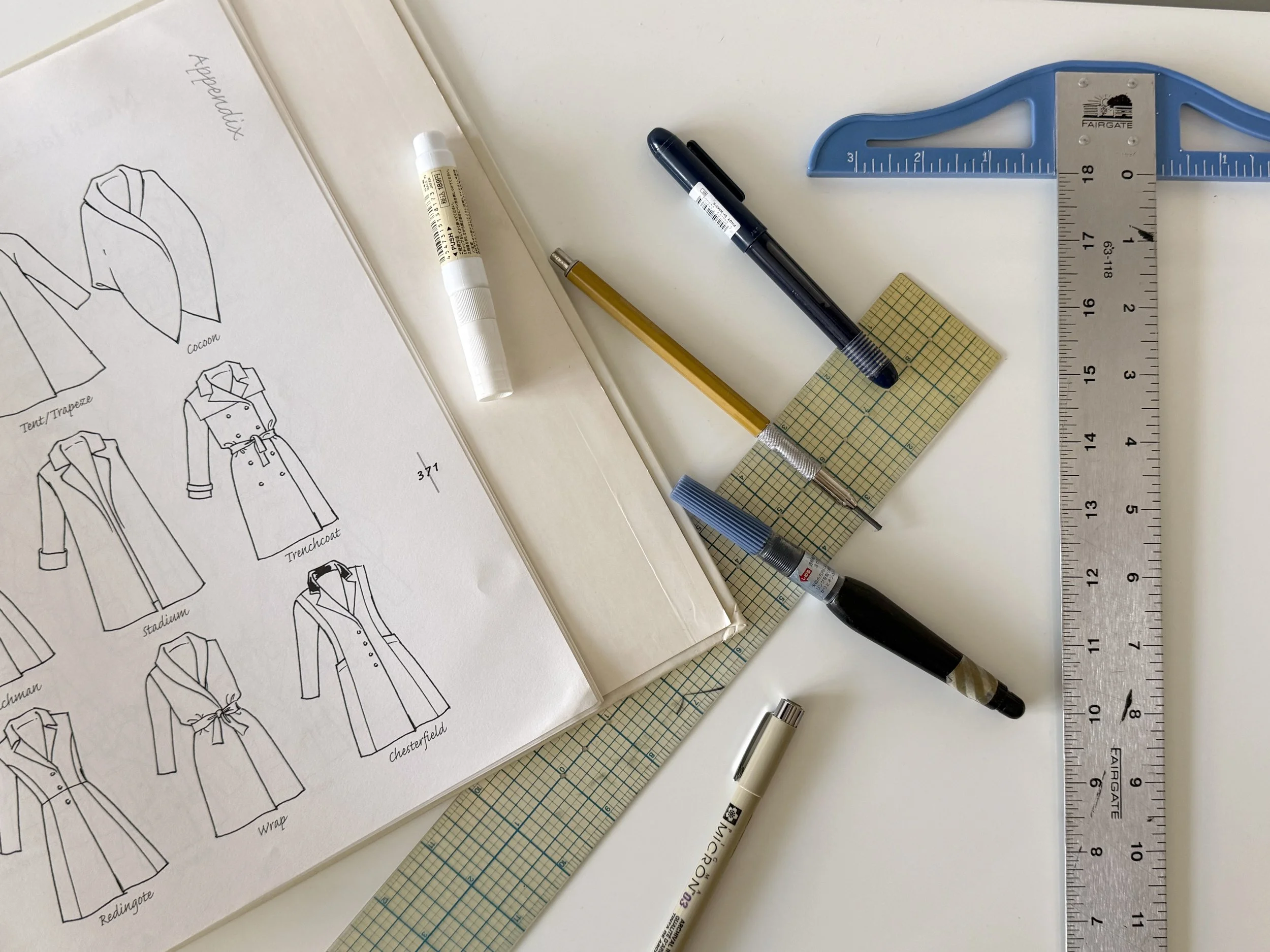 A fashion design workspace with a pattern book showing clothing illustrations, various pens, a ruler, a cutting mat, and a large blue square ruler on a white surface.