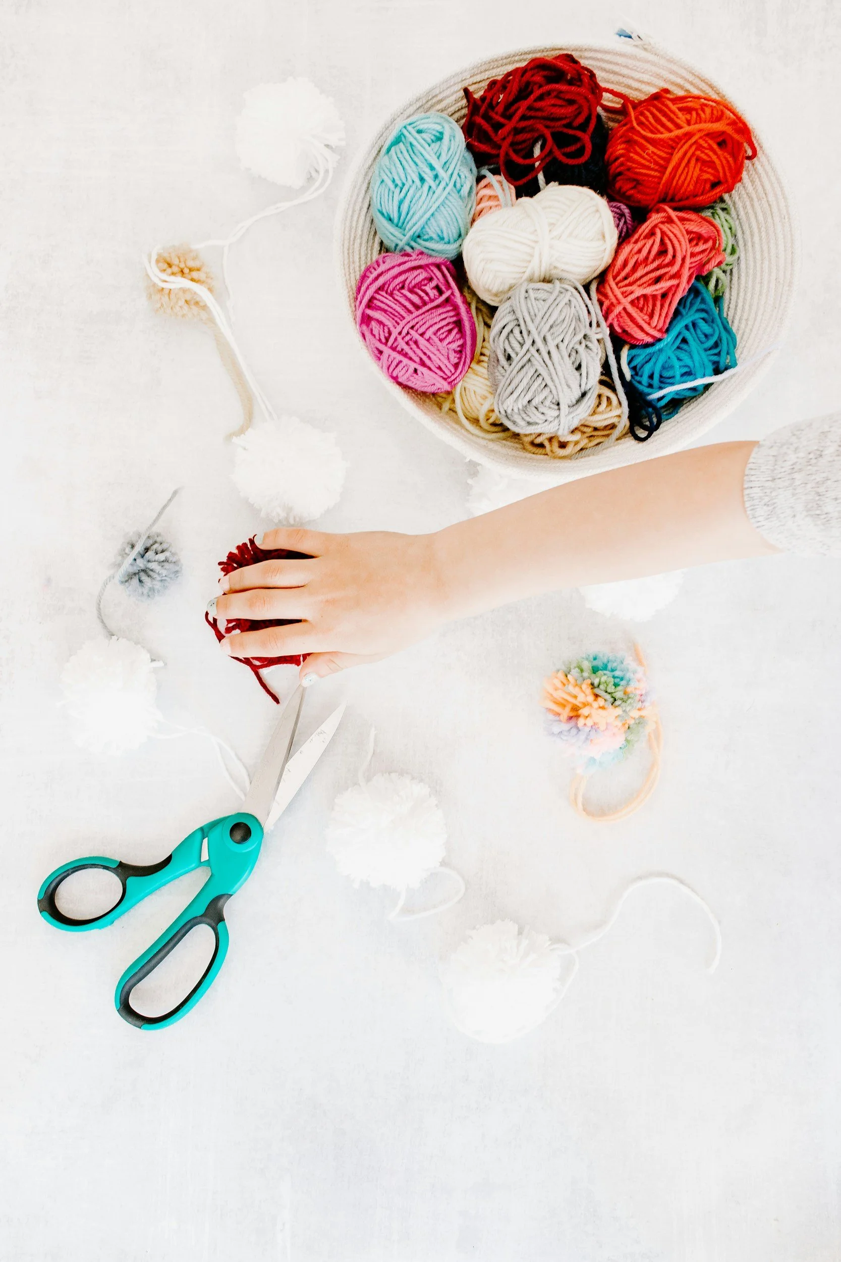 A workspace with a pair of teal and black scissors, a person's hand, and a basket of colorful yarn balls on a white surface with pom-poms and a crochet hook.