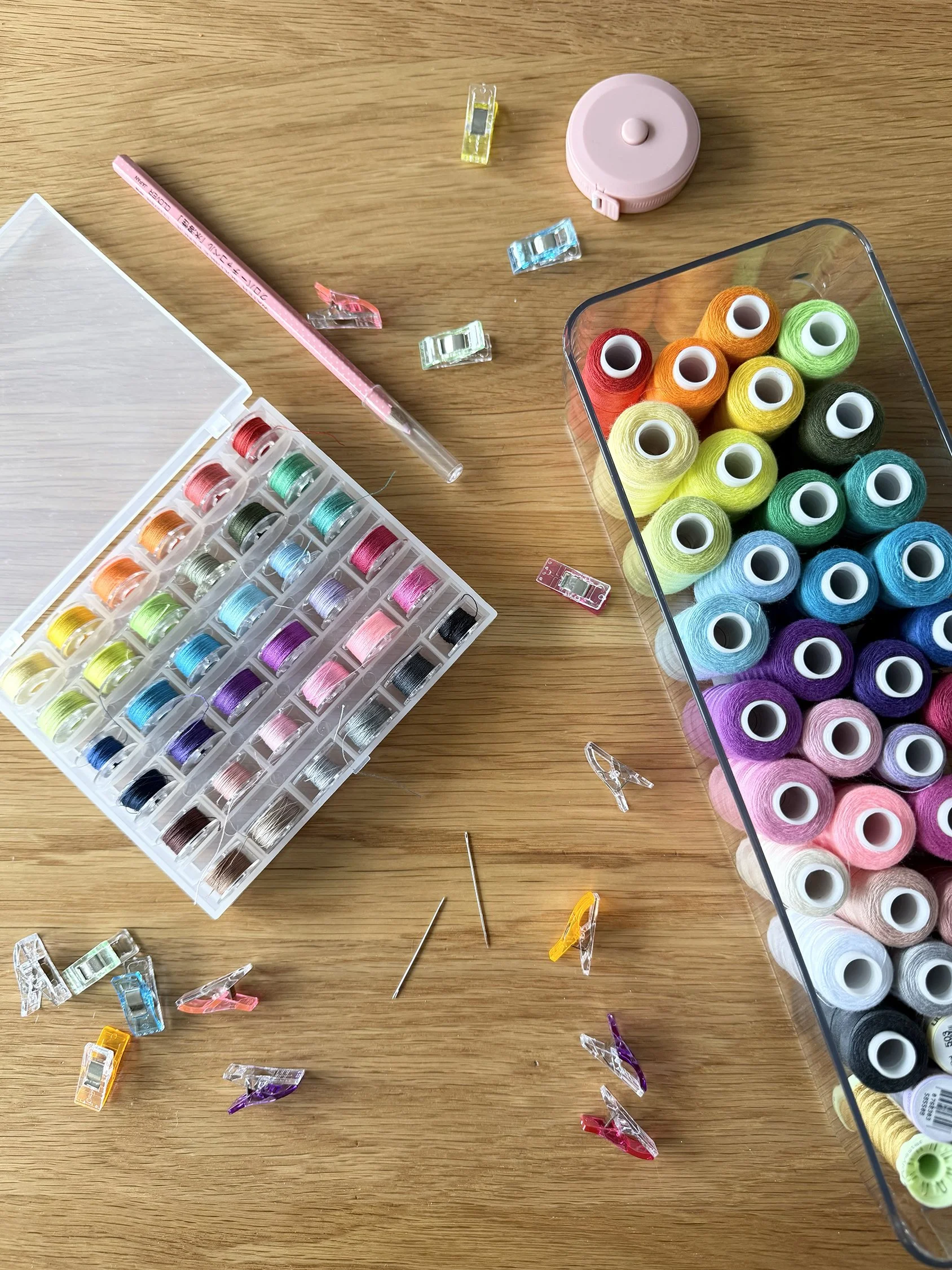 A collection of colorful sewing thread spools, empty plastic clips, a tape measure, and sewing pins on a wooden surface.