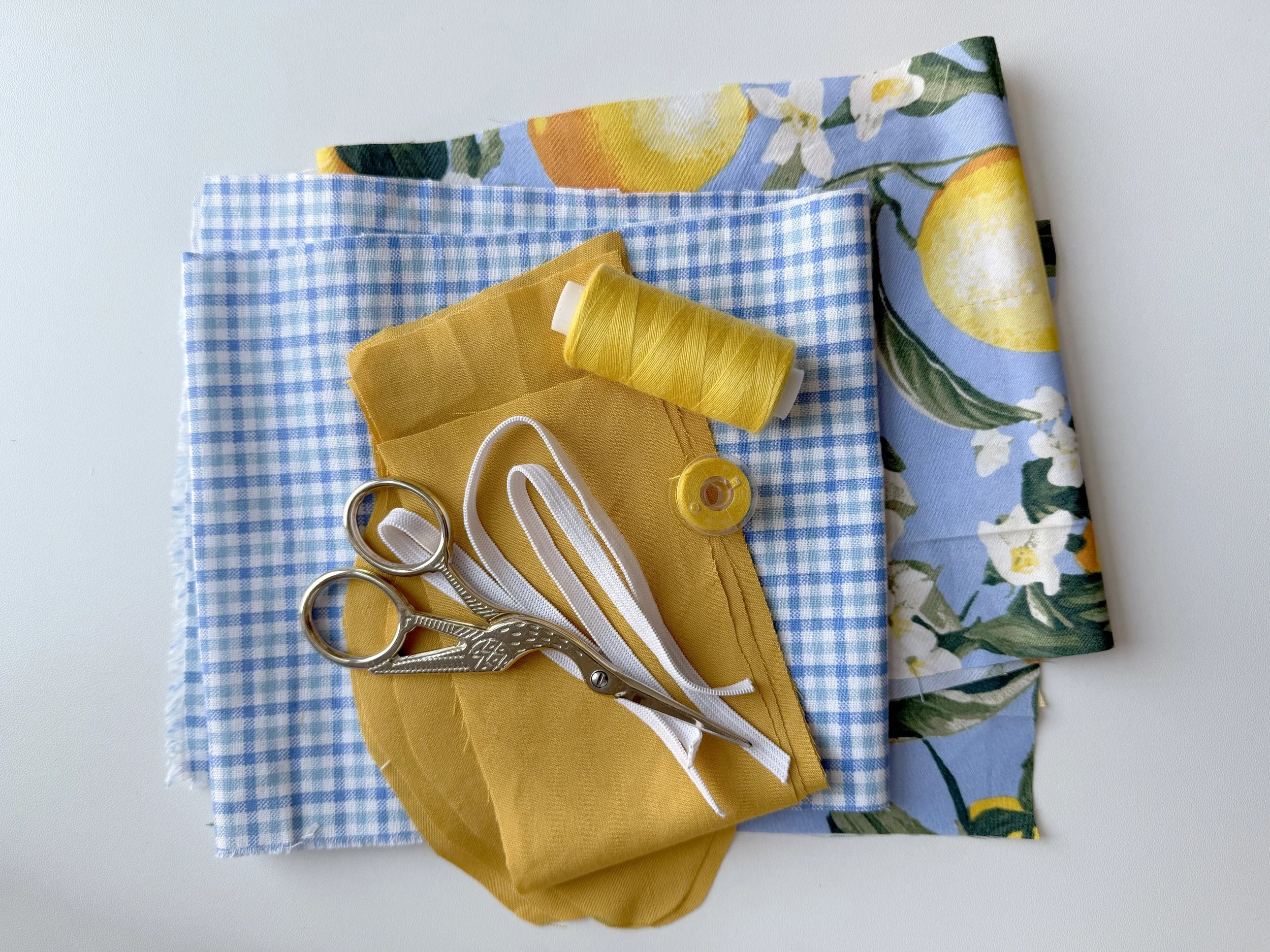 A collection of sewing and fabric items including a blue checked cloth, yellow fabric, white elastic bands, yellow spool of thread, silver scissors, and floral fabric with lemons and white flowers, arranged on a white surface.
