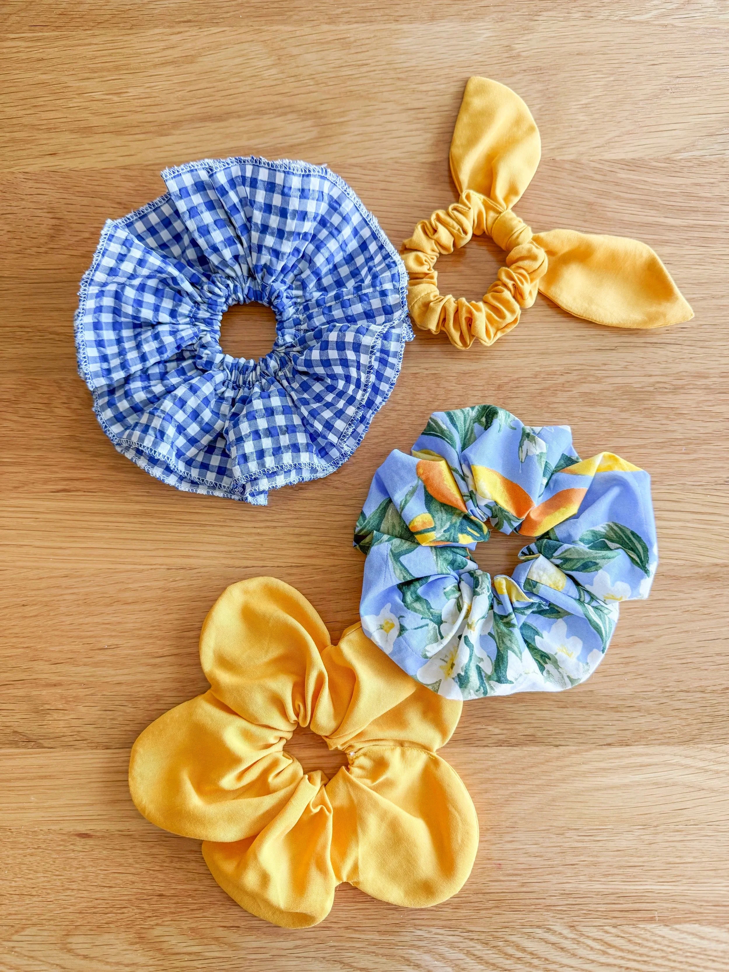 Three fabric scrunchies on a wooden surface: one blue and white gingham, one yellow satin, and one floral with green, yellow, and orange patterns.