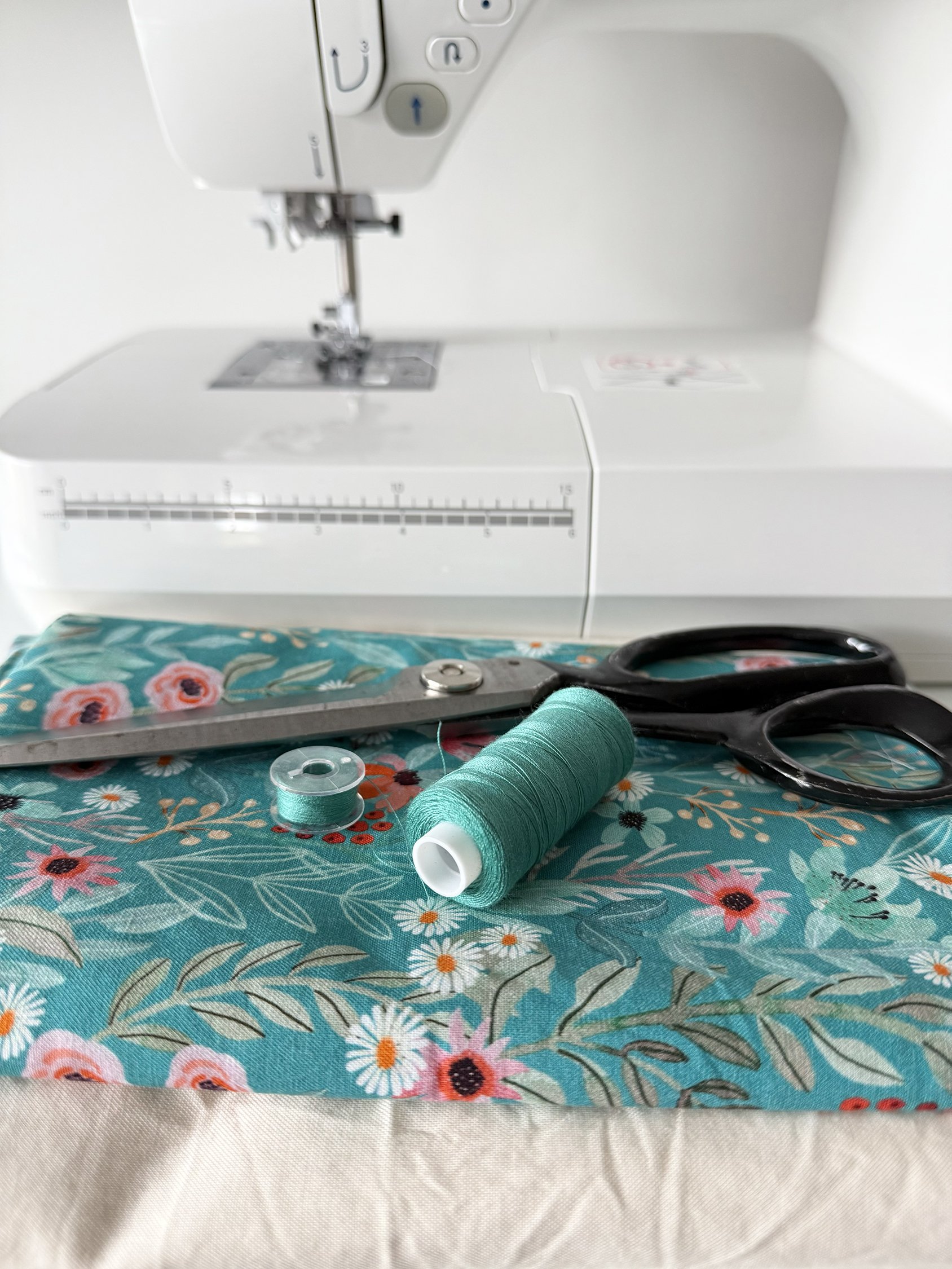 A sewing machine with floral fabric underneath, red scissors, and sewing supplies including a spool of teal thread and a small button.