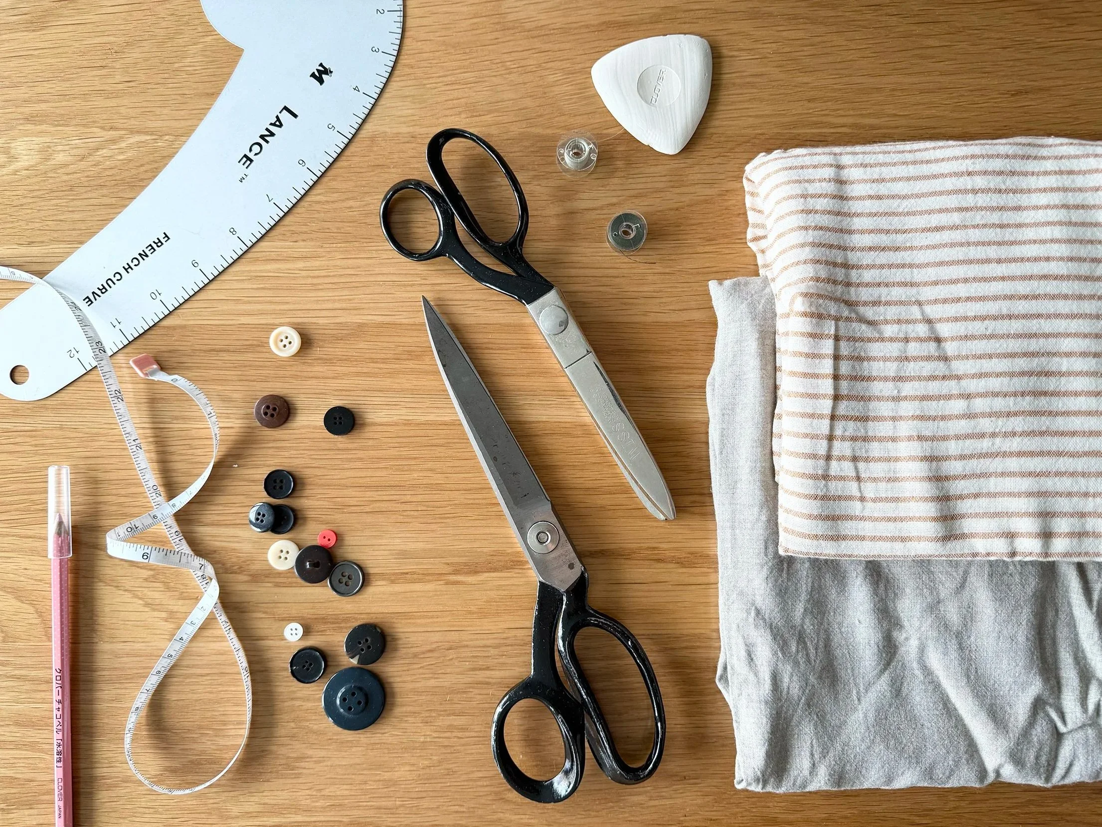 A wooden table with sewing tools and materials, including a measuring tape, a pair of scissors, thread spools, buttons, a fabric piece, a heart-shaped object, and a small plastic container.