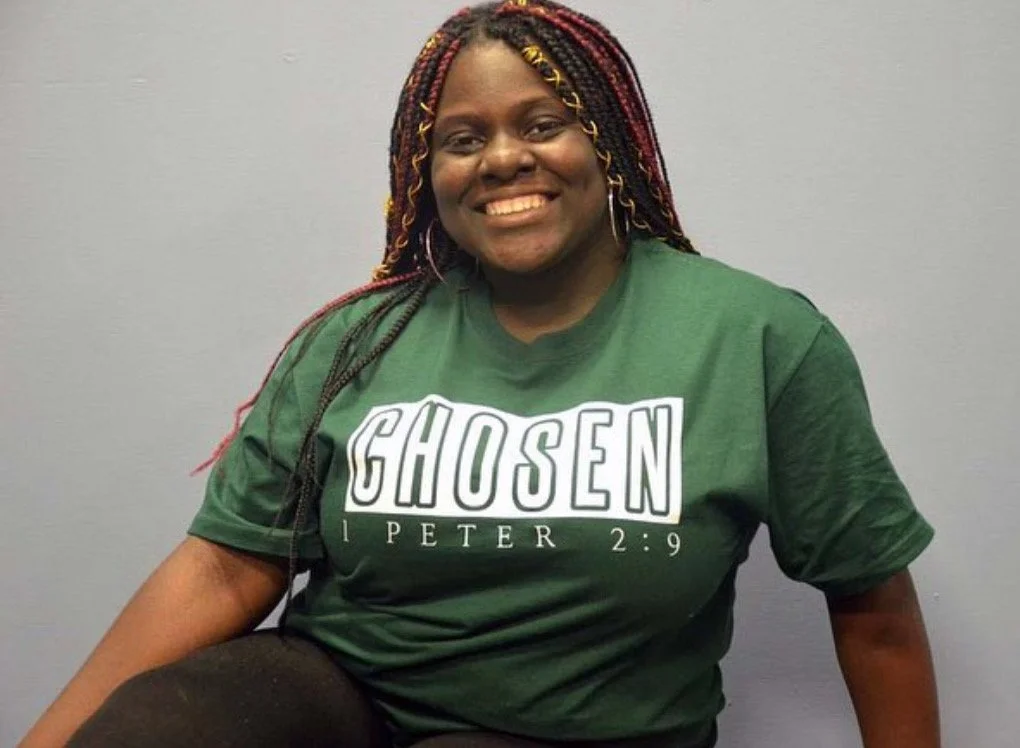 A woman with braided hair wearing a green T-shirt with the word 'CHOSEN' and a Bible verse, smiling at the camera.